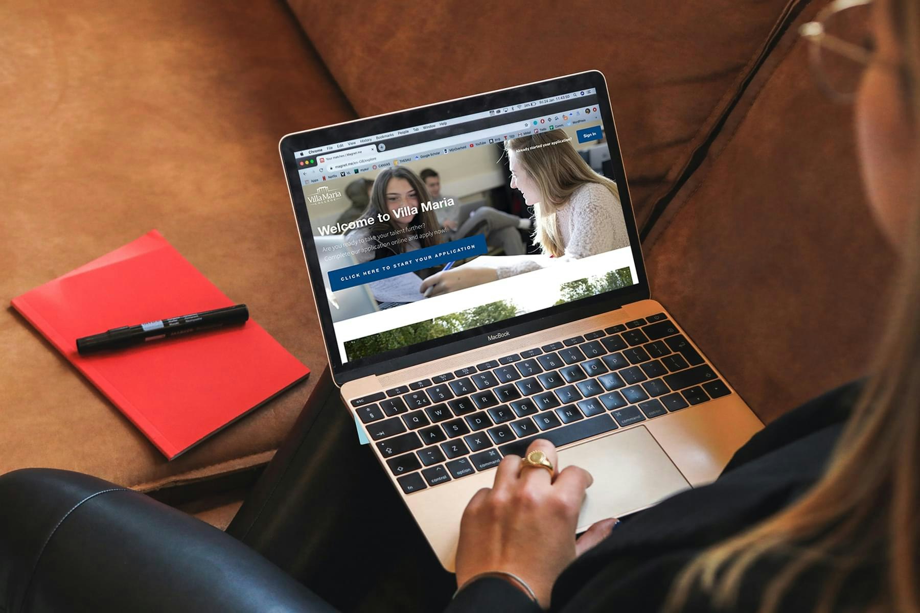 Picture of a woman browsing the Villa Maria website on her laptop, and having a red notebook next to her on the sofa.