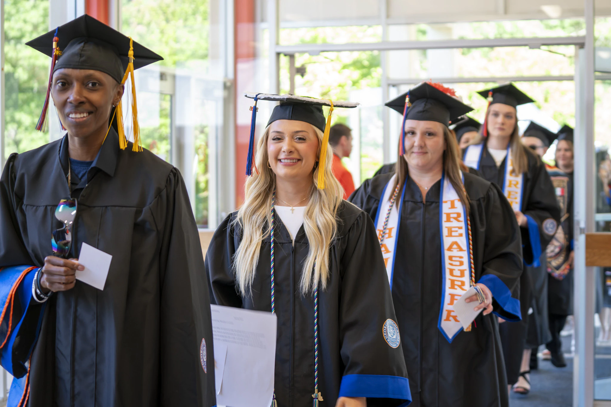 Smiling Georgia Highlands College graduates in caps and gowns walking in a commencement processional