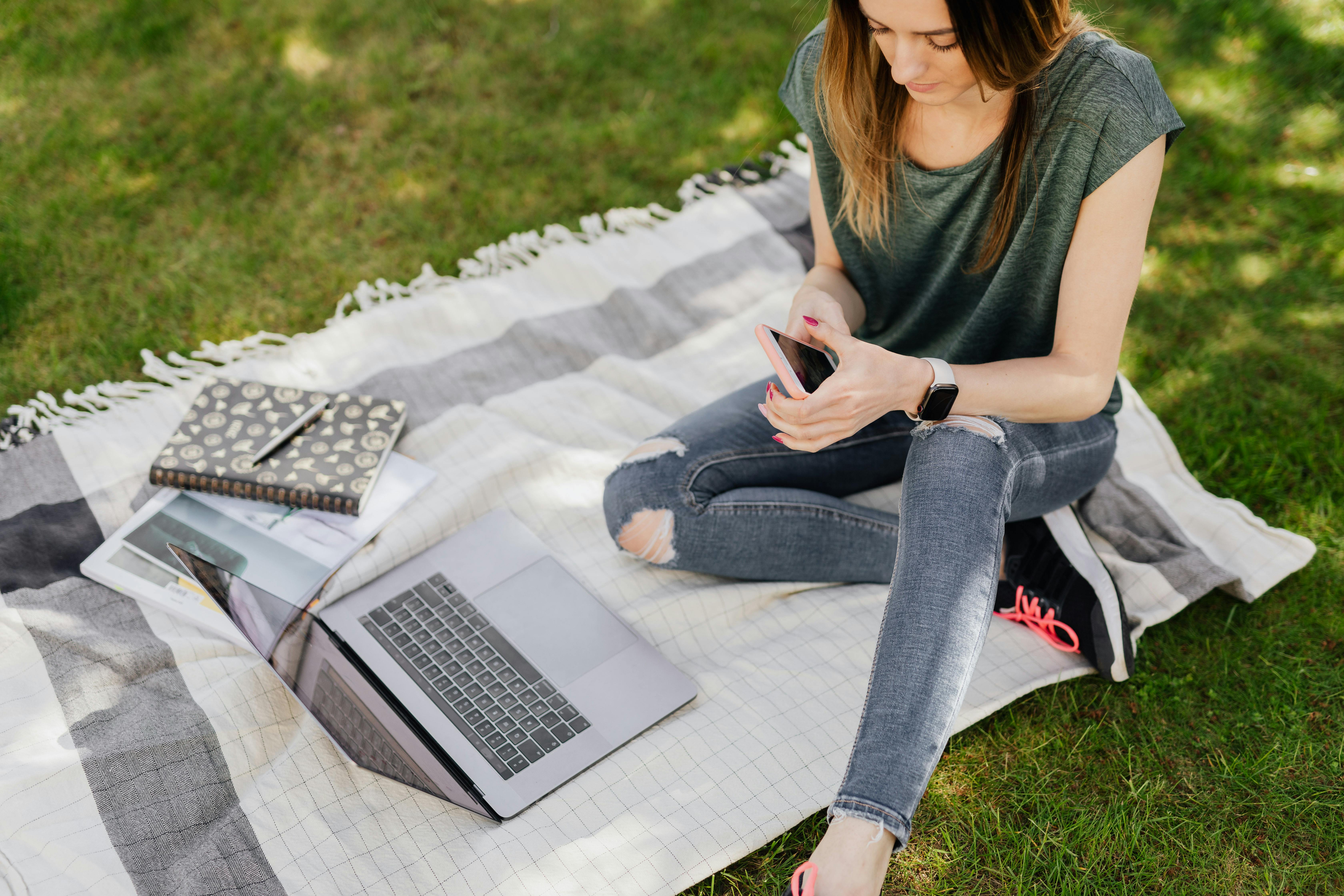 Female student texting on a phone with a laptop and books outside