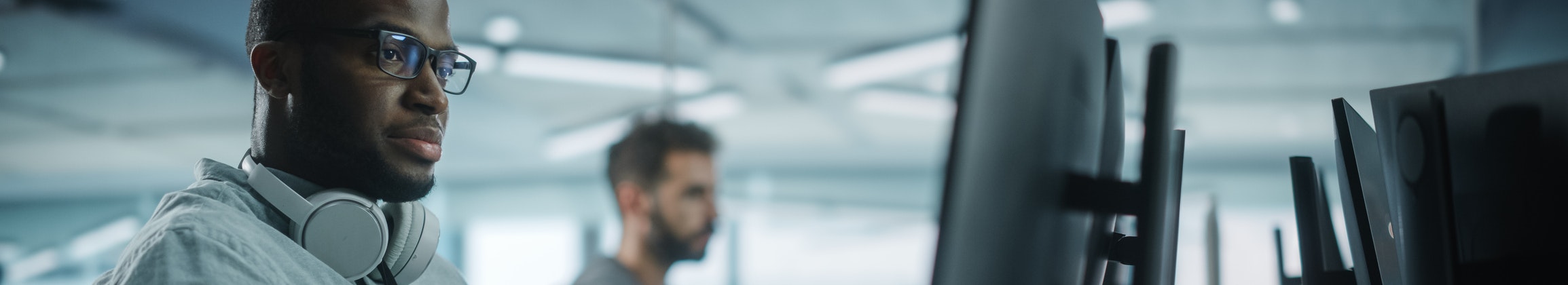 This image shows two men working at computer desks in a modern, open office setting. The man in the foreground is wearing glasses, a light gray shirt, and has a pair of large white headphones resting around his neck. He is focused on a task, typing on a keyboard while looking at a large monitor. In the background, another man is also working at a computer. The workspace is filled with multiple monitors and cables, and the lighting is soft and natural, giving the scene a calm, focused atmosphere.