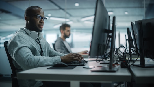This image shows two men working at computer desks in a modern, open office setting. The man in the foreground is wearing glasses, a light gray shirt, and has a pair of large white headphones resting around his neck. He is focused on a task, typing on a keyboard while looking at a large monitor. In the background, another man is also working at a computer. The workspace is filled with multiple monitors and cables, and the lighting is soft and natural, giving the scene a calm, focused atmosphere.