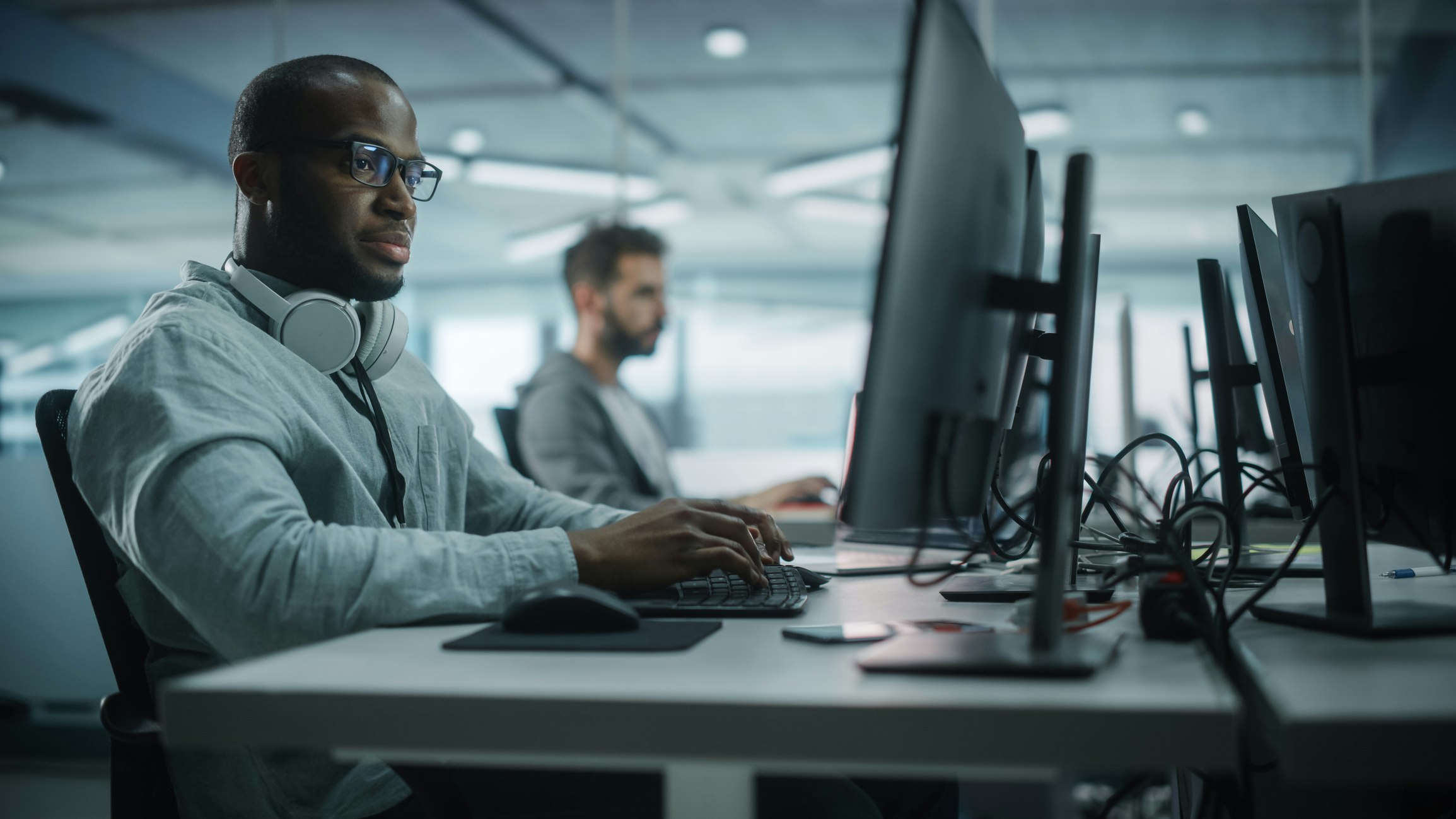 This image shows two men working at computer desks in a modern, open office setting. The man in the foreground is wearing glasses, a light gray shirt, and has a pair of large white headphones resting around his neck. He is focused on a task, typing on a keyboard while looking at a large monitor. In the background, another man is also working at a computer. The workspace is filled with multiple monitors and cables, and the lighting is soft and natural, giving the scene a calm, focused atmosphere.