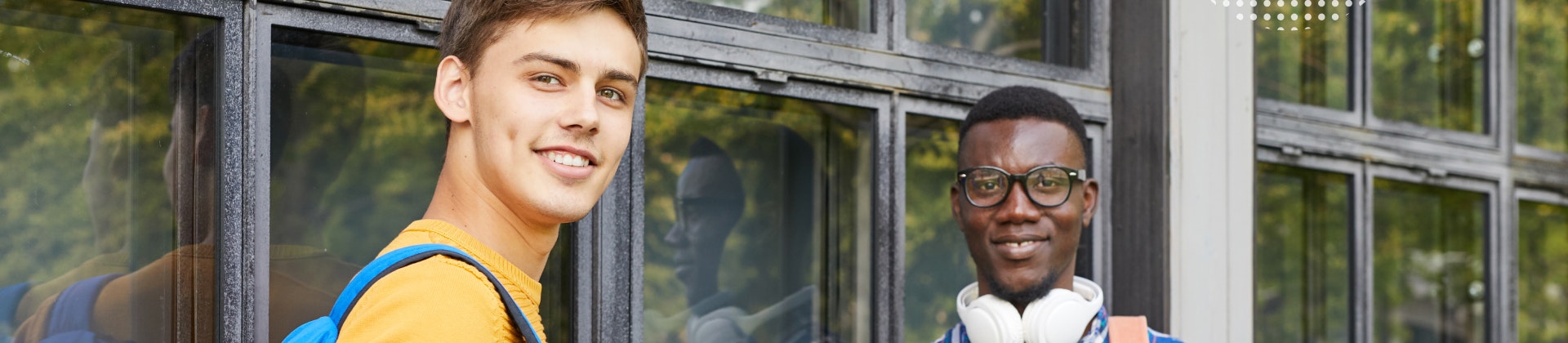 A white male student and a Black male student leaning up against windows wearing backpacks and holding notebooks and laptops.