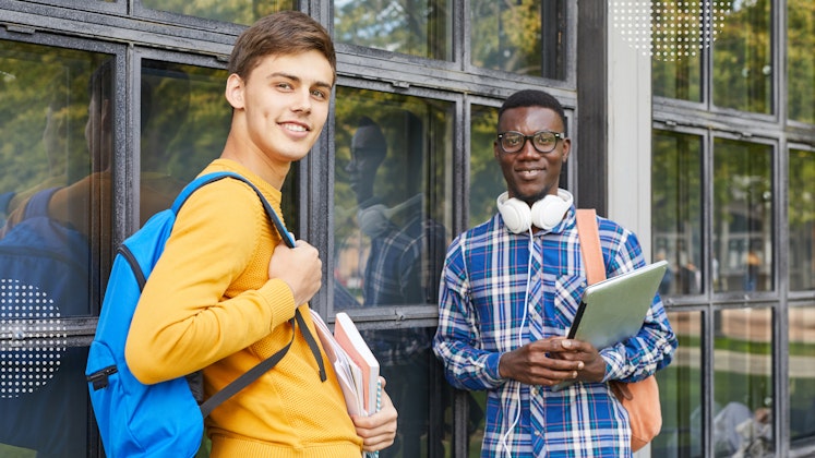 A white male student and a Black male student leaning up against windows wearing backpacks and holding notebooks and laptops.