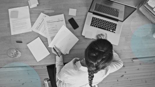 A girl is working on a laptop with scattered papers, a cell phone, and a glass of water next to her.