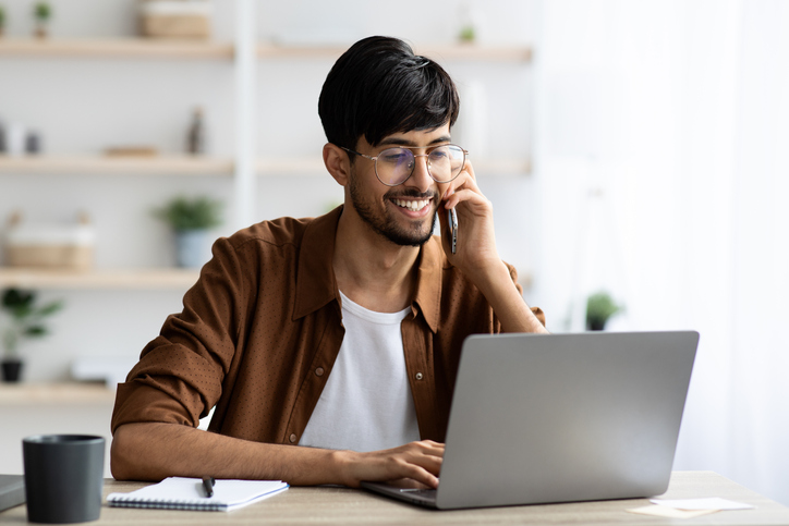 Cheerful businessman sitting at workdesk with laptop having phone conversation with business partner or client, typing on computer keyboard, office interior, copy space