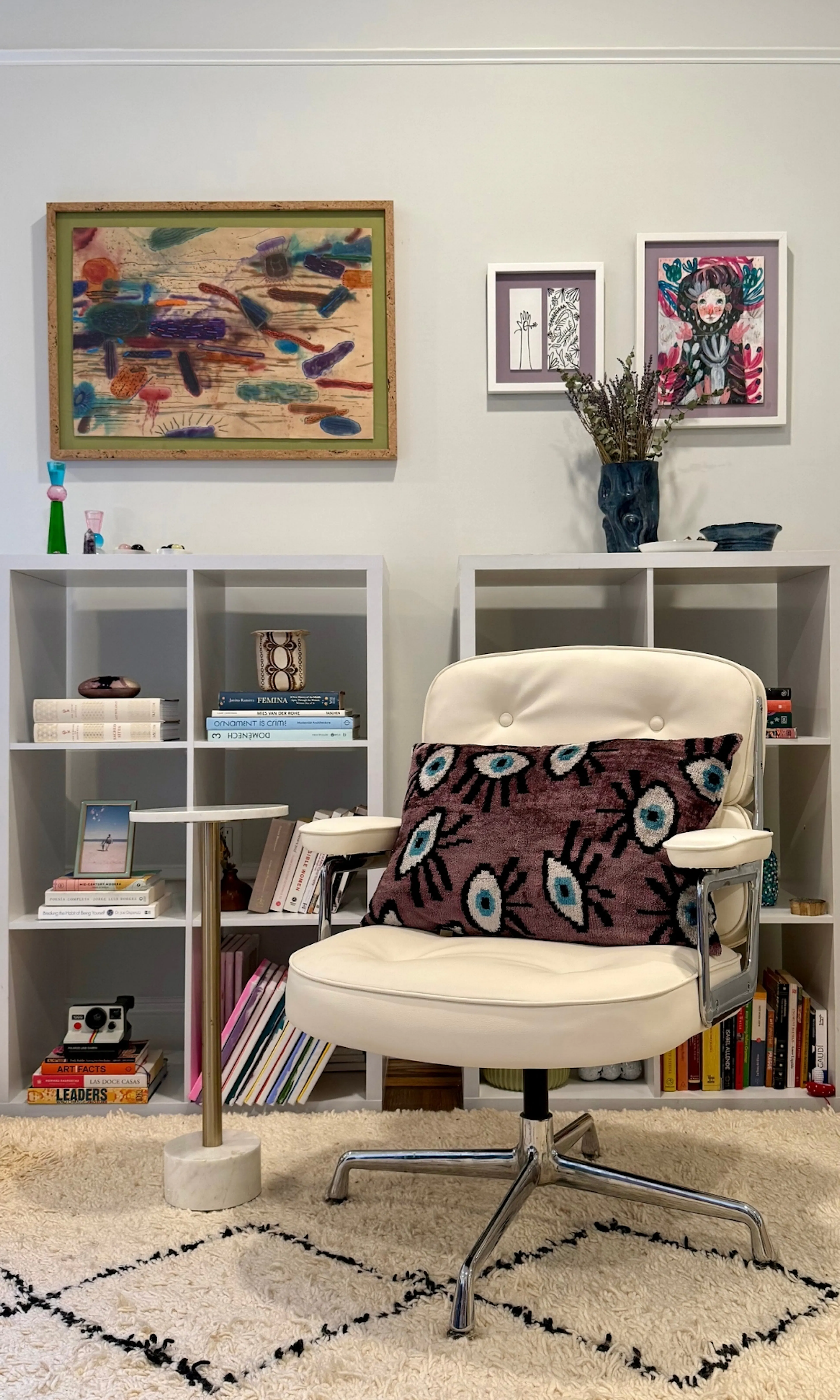an image of an eames chair with a purple pillow in front of a white bookcase with books and objects as well as art on the wall.