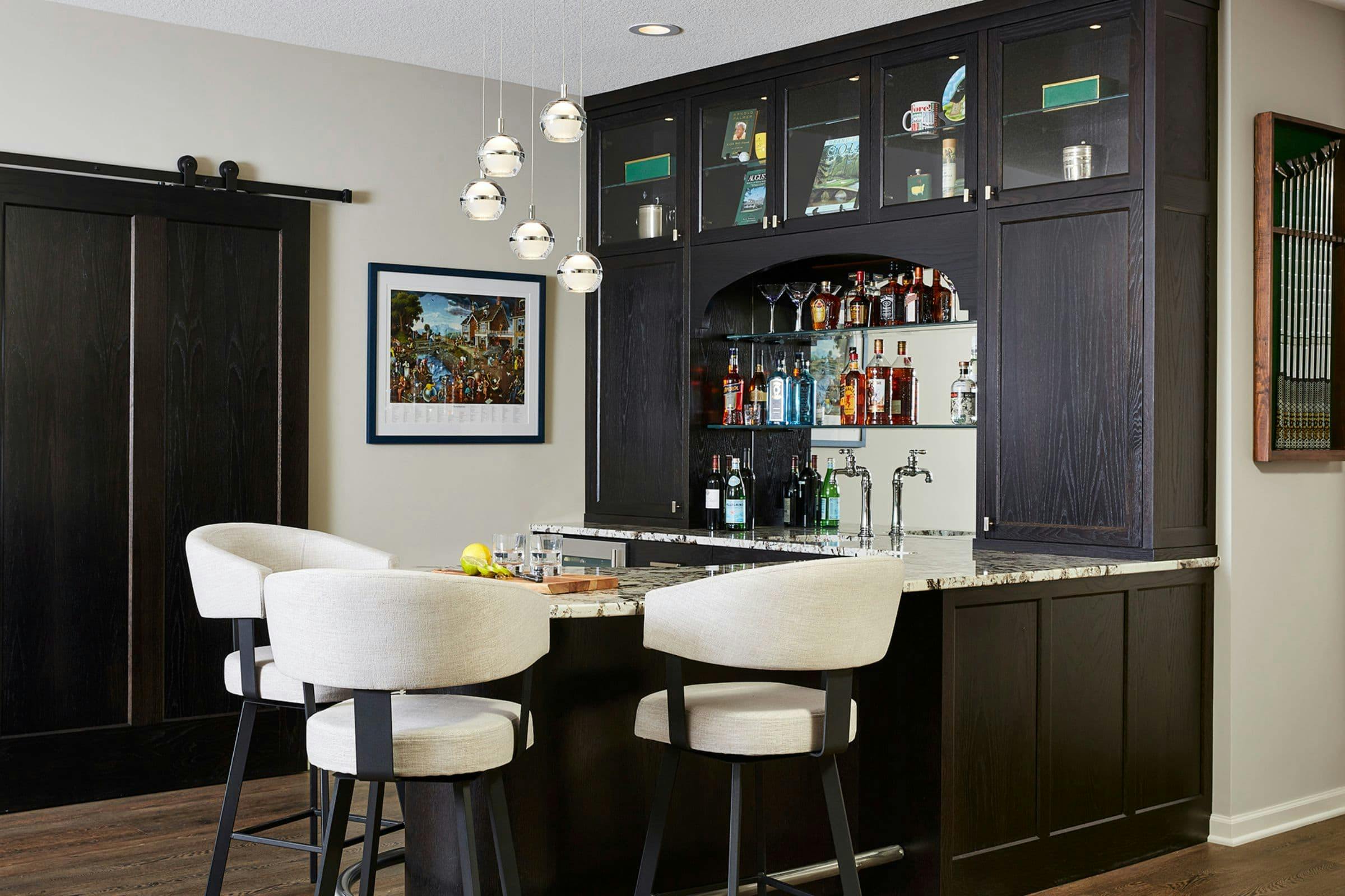 dark cabinetry with paneled front used in an elegant lower level home bar design with seating and light fixture