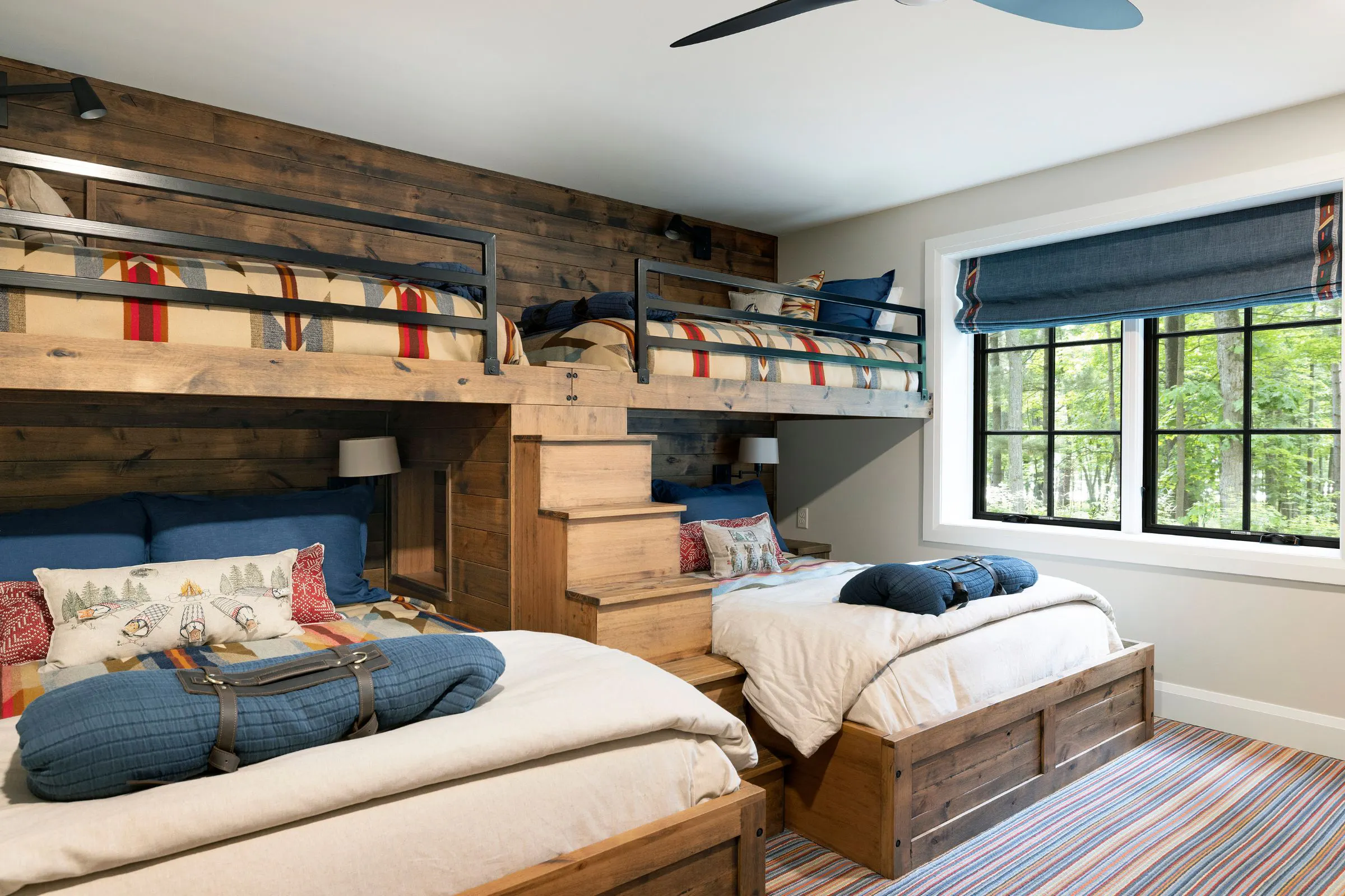 Bunk room with built-in wood bunk beds, blue bedding, colorful striped carpet and black-framed windows overlooking the woods.
