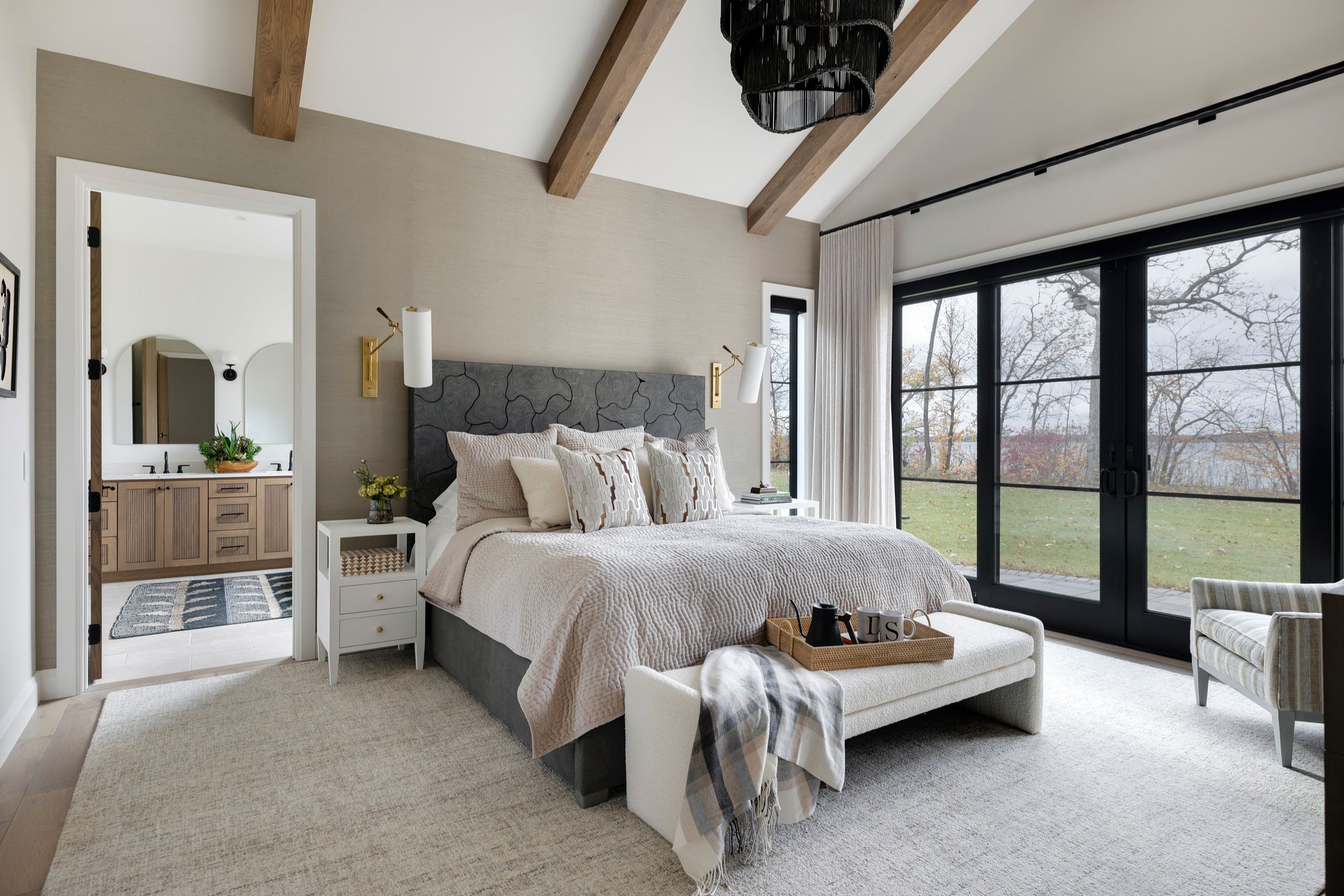 A warm, inviting primary bedroom featuring a vaulted ceiling with exposed wood beams, layered neutral textures, and a sculptural black pendant.