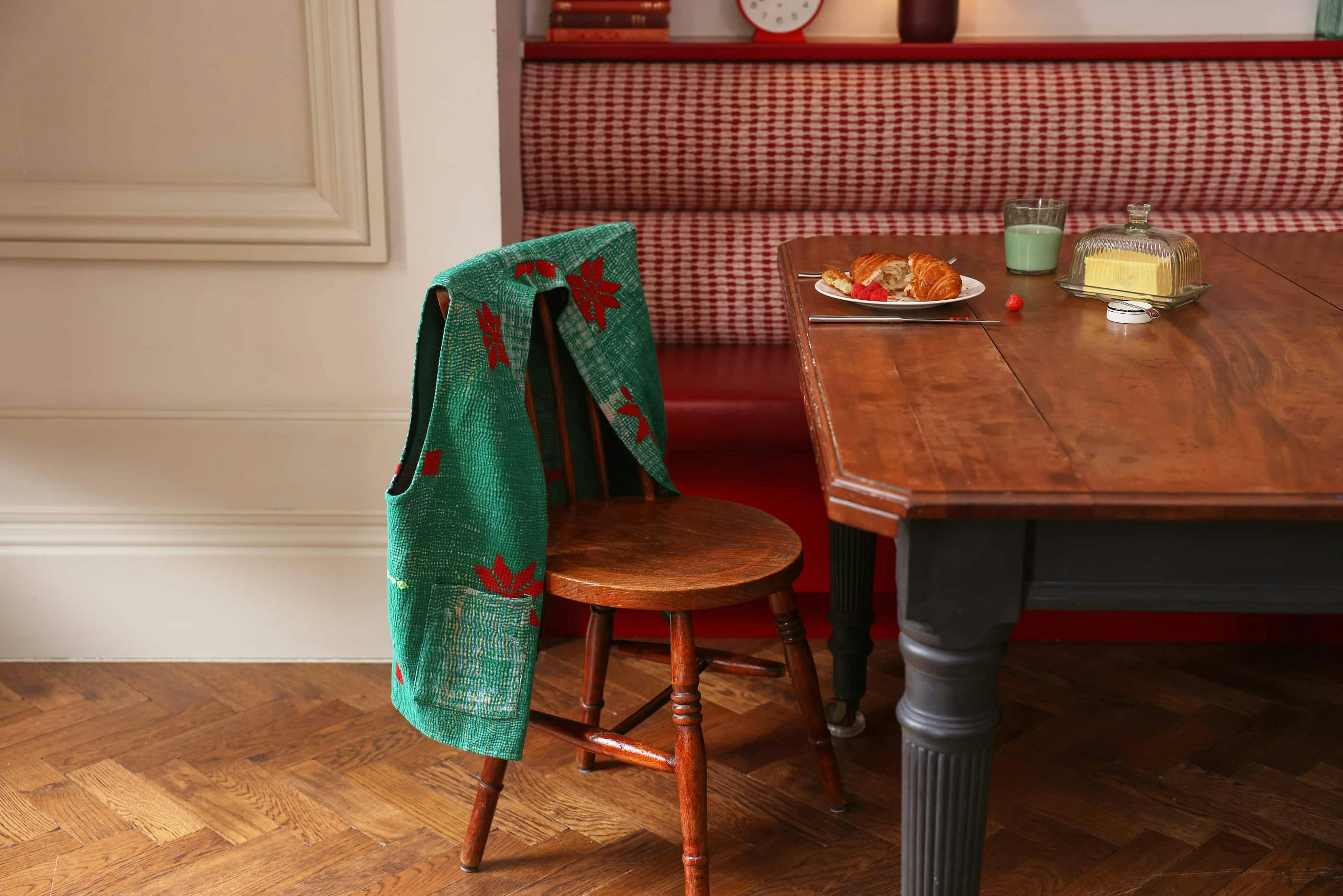 An Indigo Wares green waistcoat draped over the back of a chair which is next to a table with a croissant on a plate and a butter dish.