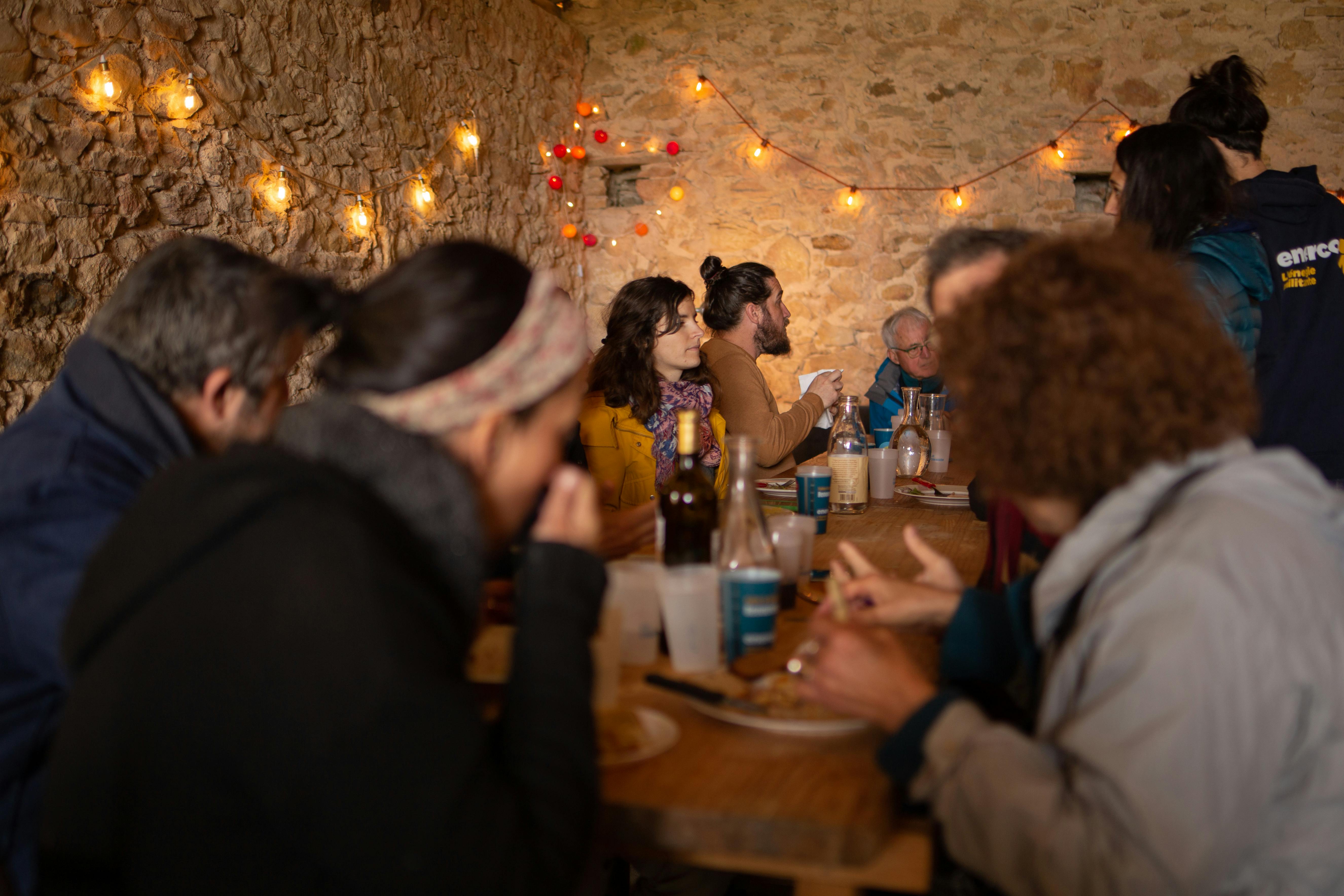 Repas avec plusieurs personnes, dans une pièce avec pierres apparentes. Guirlandes de fêtes sur les murs.