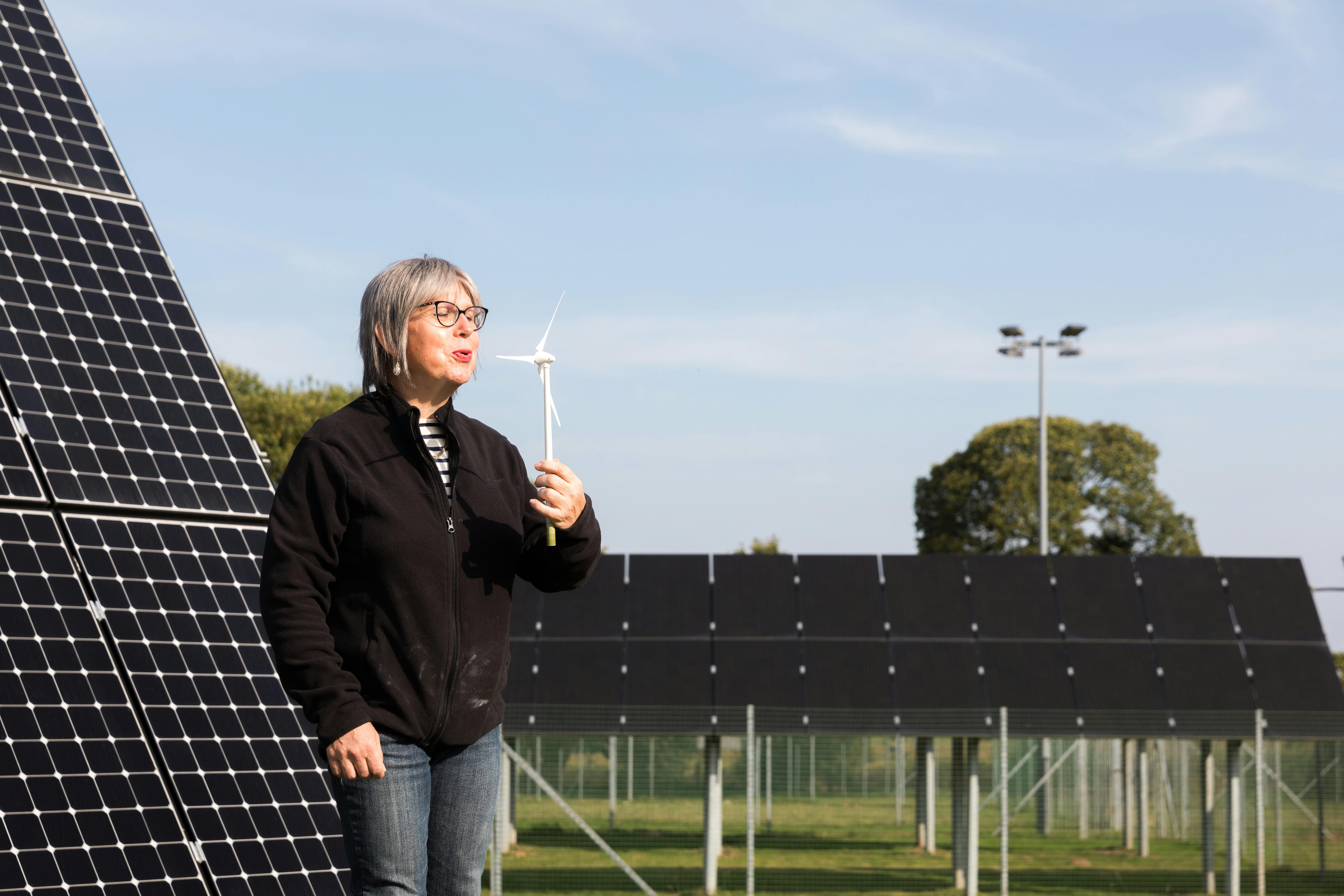 personne soufflant sur une petite éolienne, avec des panneaux photovoltaïques en fond