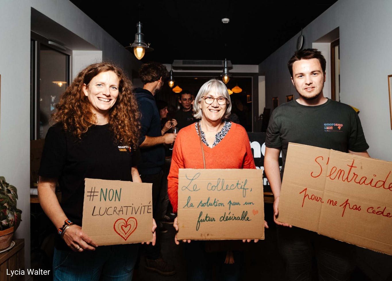 Photo de trois sociétaires d'Enercoop Nouvelle-Aquitaine avec des pancartes "#nonlucrativité", "le collectif, la solution pour un avenir désirable" et "s'entraider pour ne pas céder"