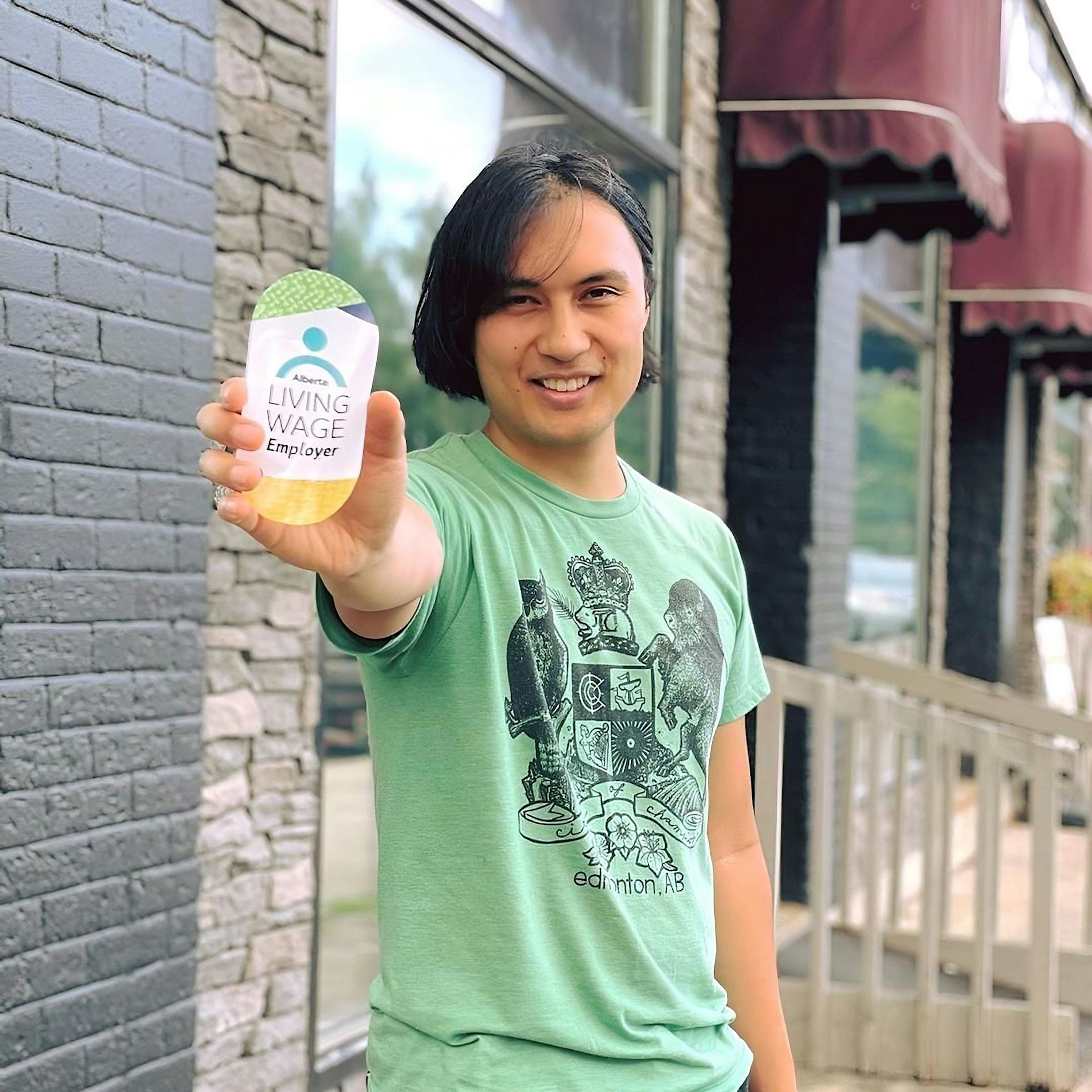 A person in a green shirt holds out an object labeled 'Alberta Living Wage Employer'