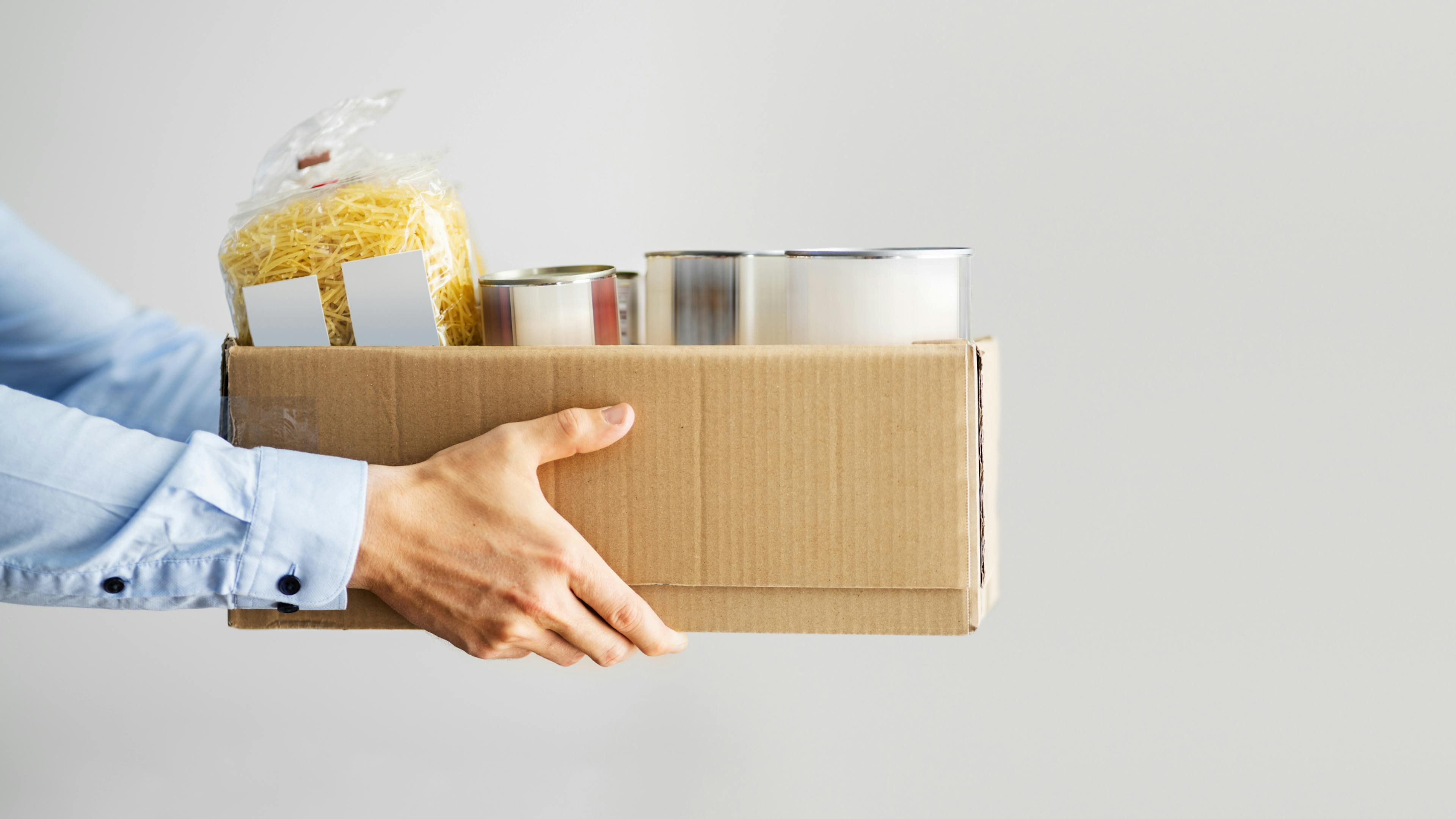 Hands holding a box of food, including canned goods.