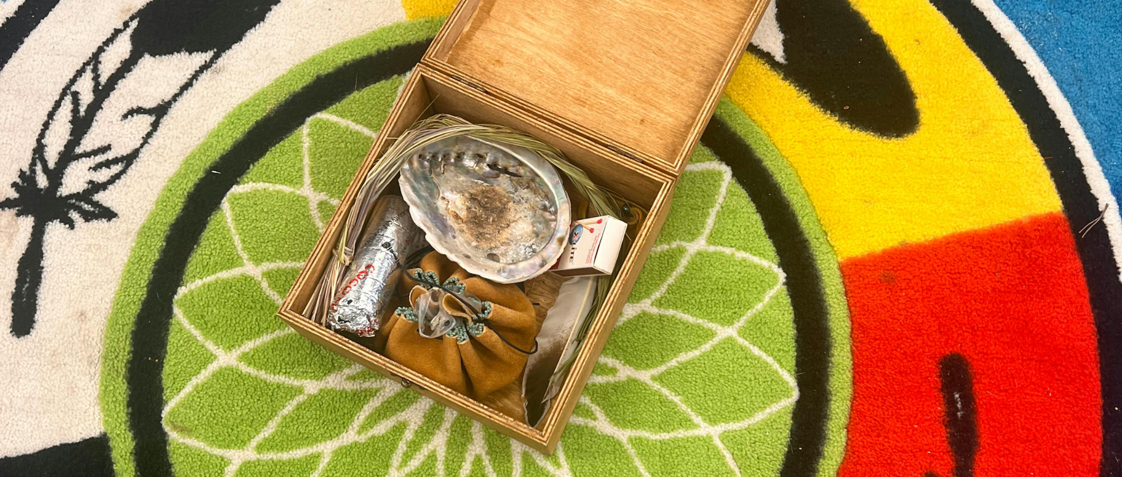 A wooden box on a colorful rug holds a shell, a pouch, and sage wrapped in foil, symbolizing Indigenous spiritual cleansing and ritual.
