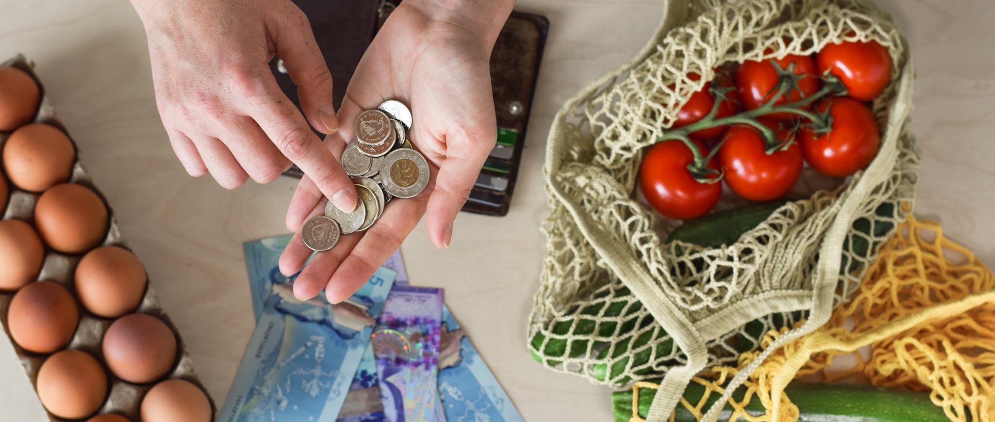 A person holds coins over a table with eggs, fresh vegetables, and cash, representing budgeting for groceries.