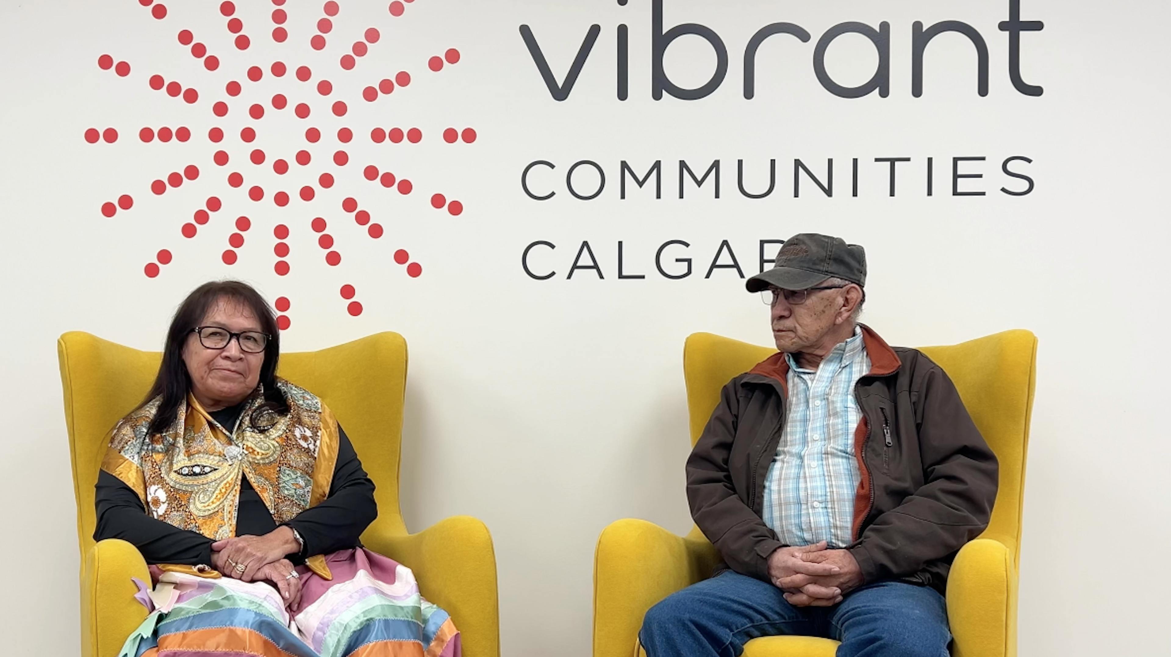 Two Elders sit in yellow chairs against a wall with "Vibrant Communities Calgary" text. One wears a colorful shawl, conveying a calm and welcoming atmosphere.
