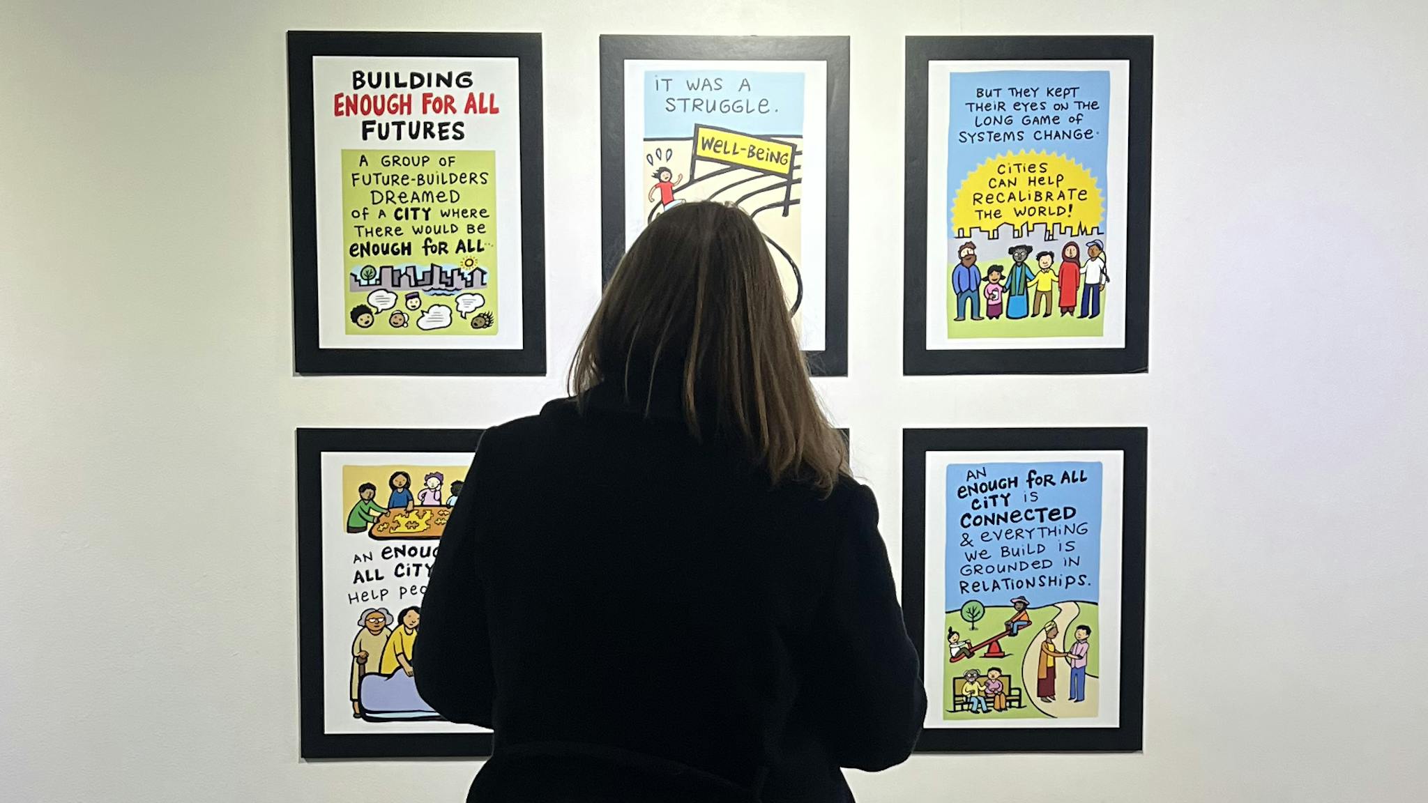 A person is standing in front of a wall featuring six zine murals depicting a community.