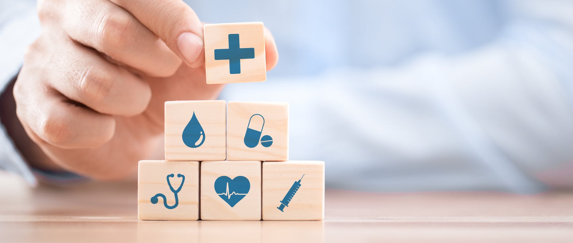 A hand stacks wooden blocks with health-themed icons: a cross, droplet, pill, stethoscope, heart, and syringe, symbolizing medical care and wellness.