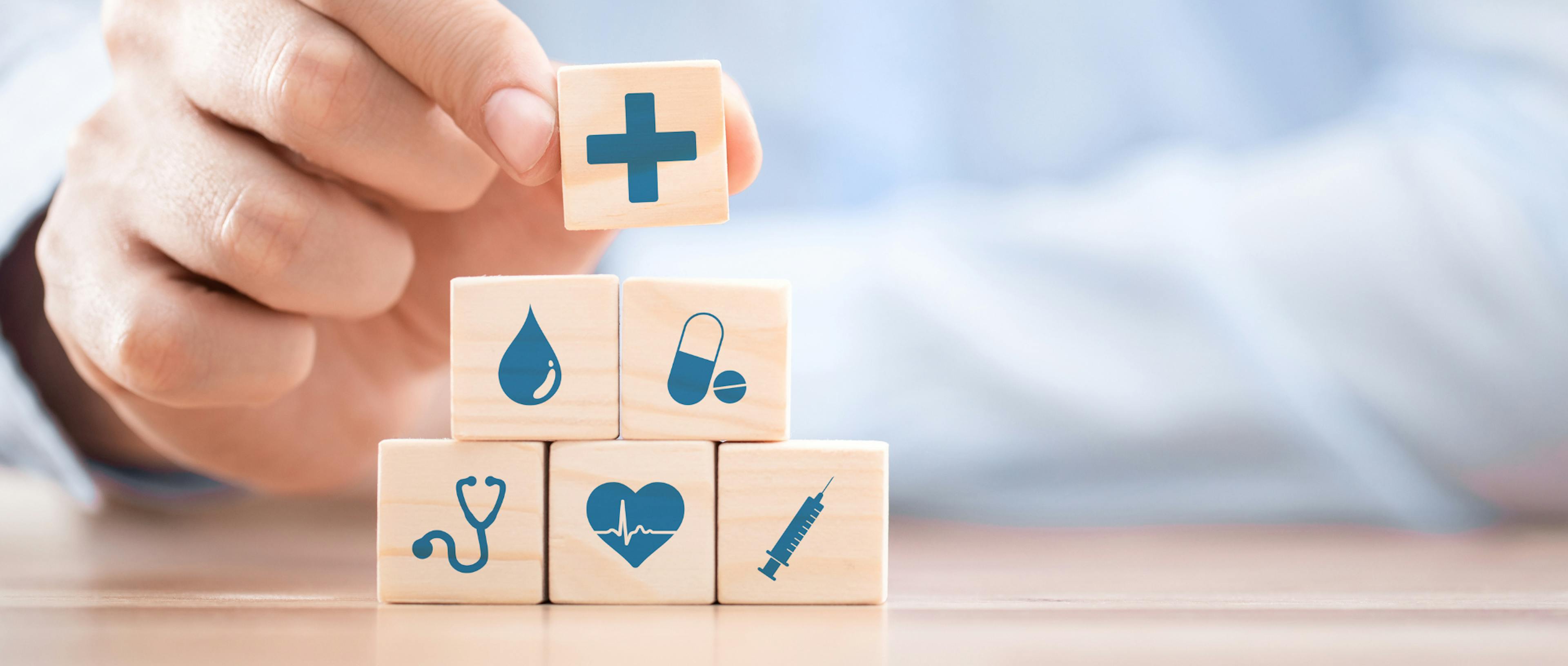A hand stacks wooden blocks with health-themed icons: a cross, droplet, pill, stethoscope, heart, and syringe, symbolizing medical care and wellness.