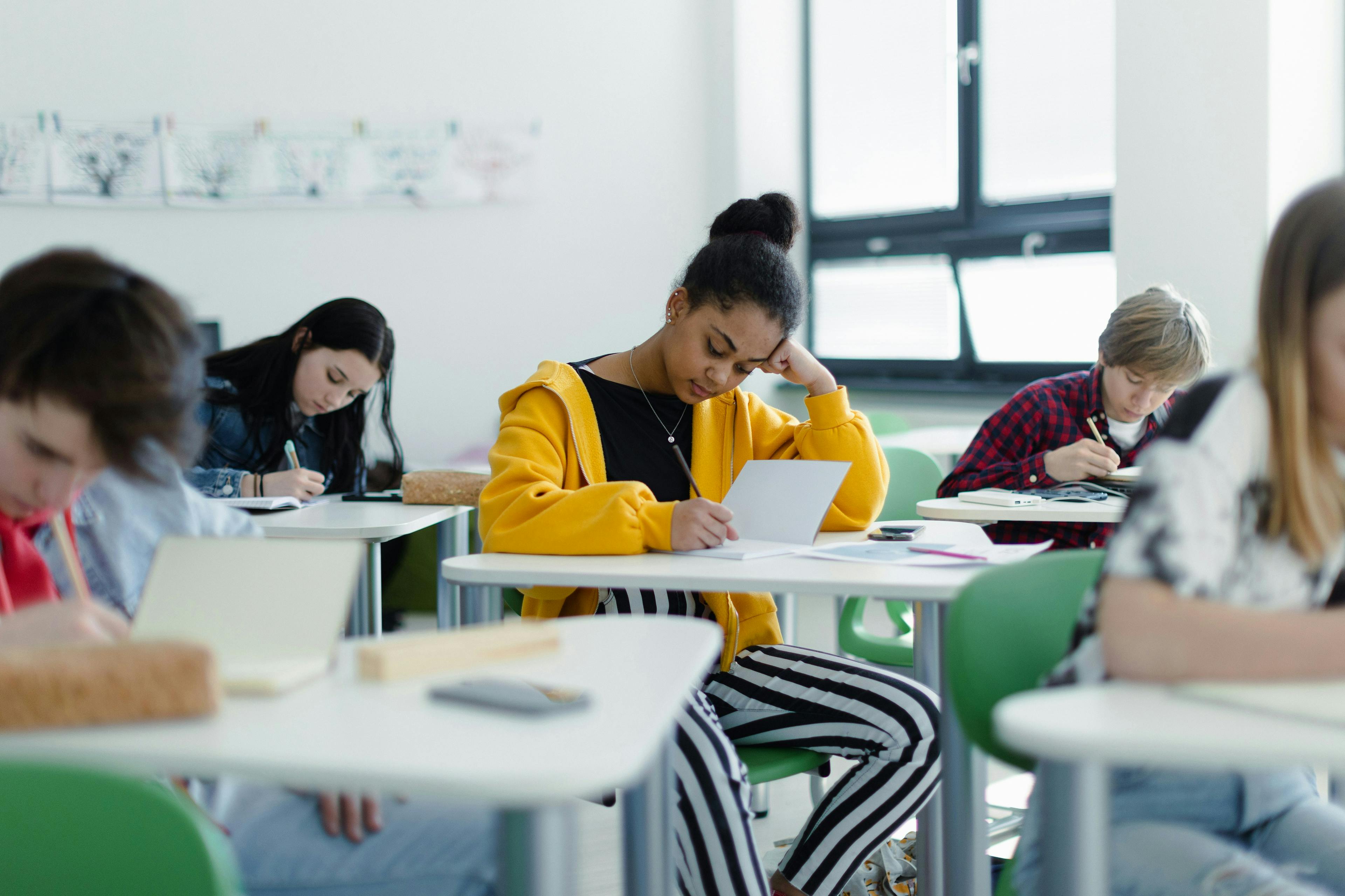 A high school student sits at her desk in the center of a classroom. There are two male students on her left and right and a female student behind. The student in the centre is resting her hand on her head as she is writing on a piece of paper on her desk. She appears to be concentrated on her work. High school completion rates trend downward in latest poverty data.