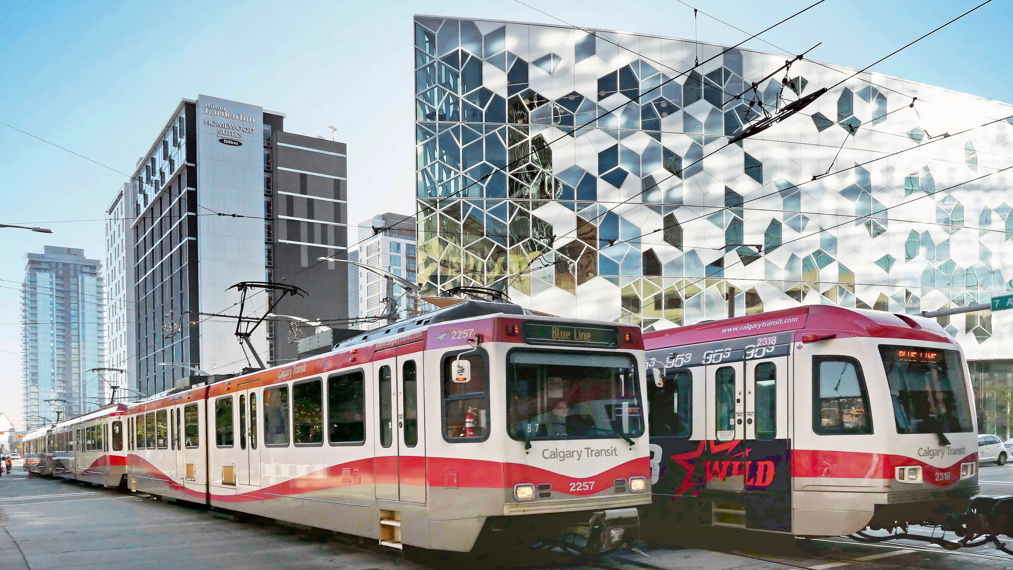 Two C-train vehicles are on the tracks in front of the Calgary Central Library