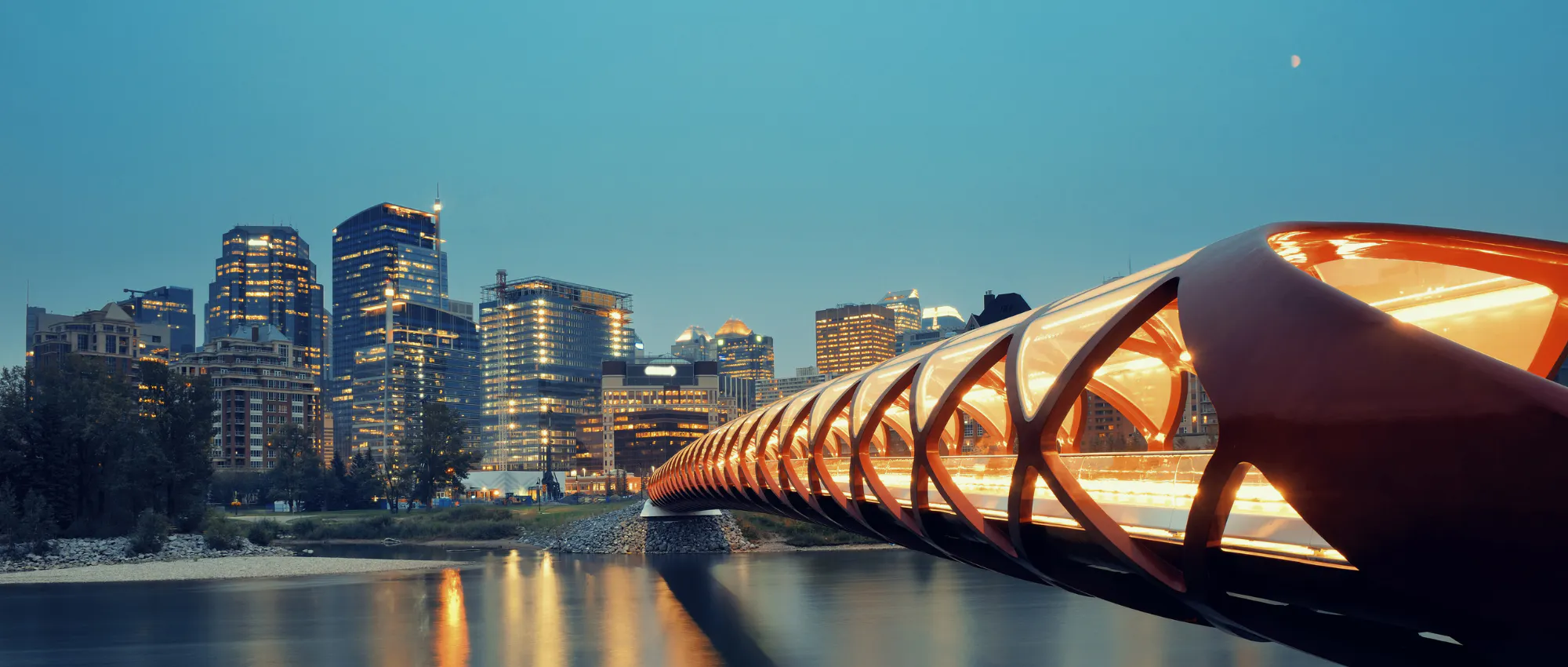 A photo of the Calgary downtown buildings featuring the peace bridge. The peace bridge is in the right side of the photo. The sky is dark, it appears to be evening time and the peace bridge is lit up, reflecting on the water below.