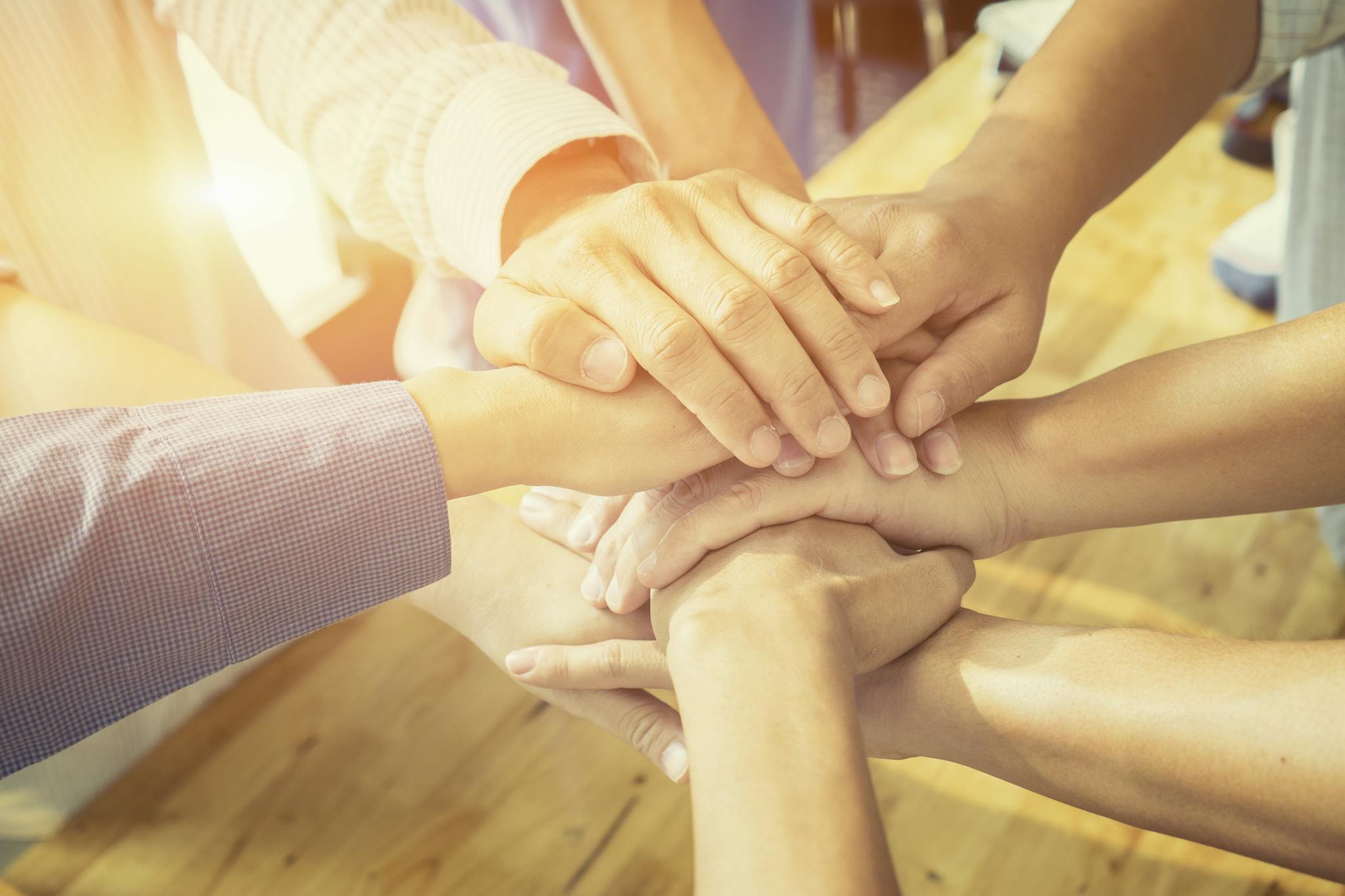 An image of six hands on top of each other in a circle. The image is bright and sunny.