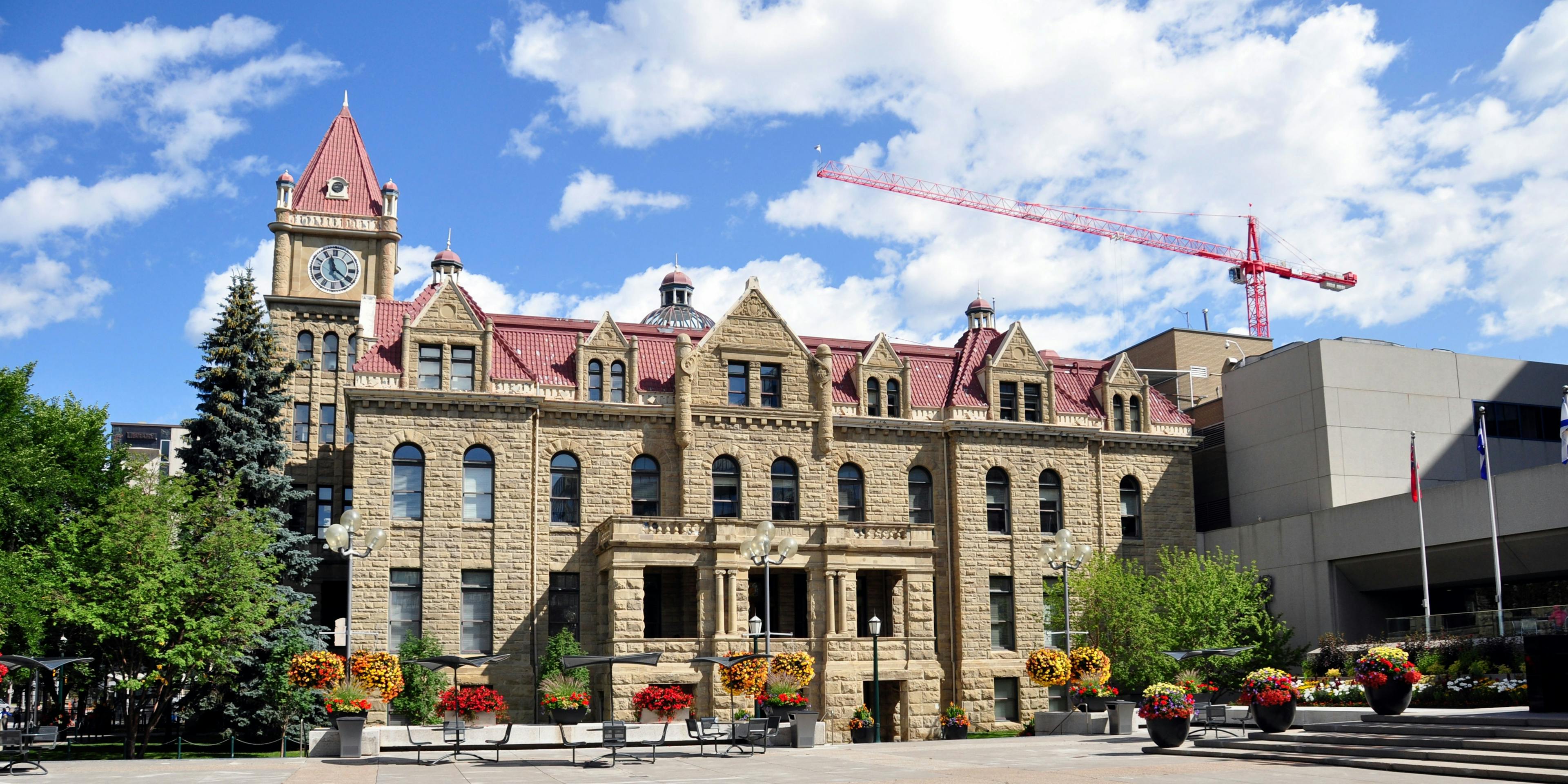 Exterior photo of Calgary City Hall Heritage Building with a blue cloudy sky and green trees and flower baskets with red and yellow flowers