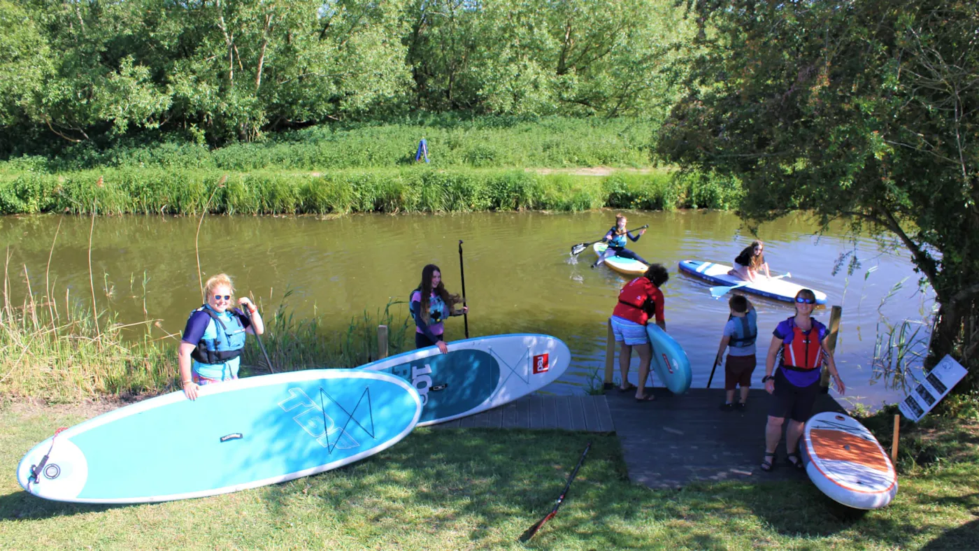 A group with stand up paddleboards at a riverside launch point on the River Stort, with others already on the water