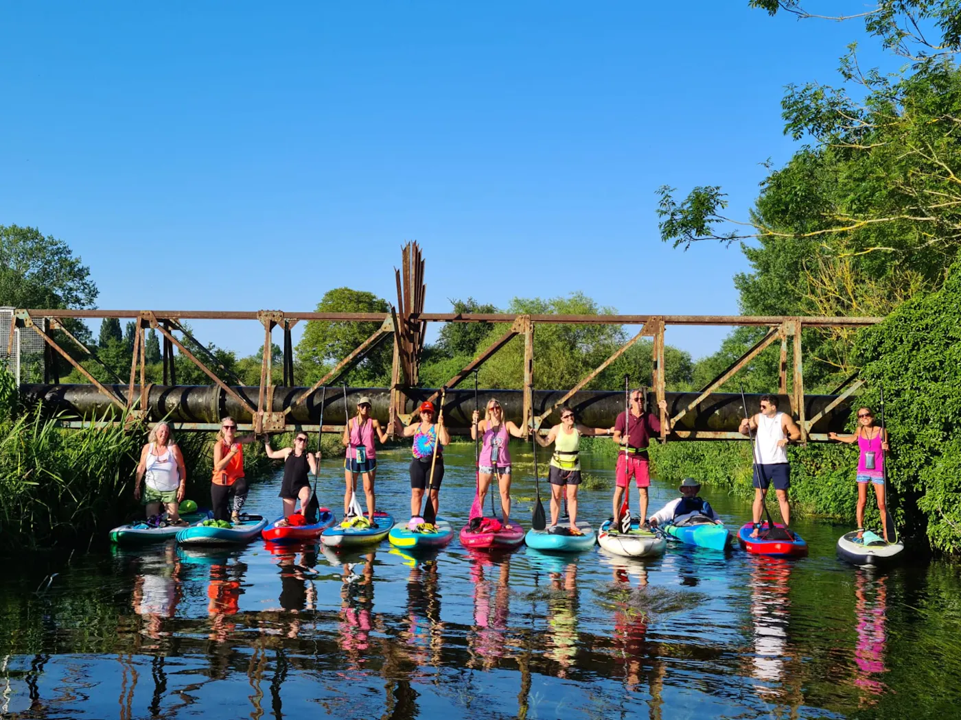 A group of paddleboarders standing on their boards under an old iron bridge on the River Medway in Tonbridge