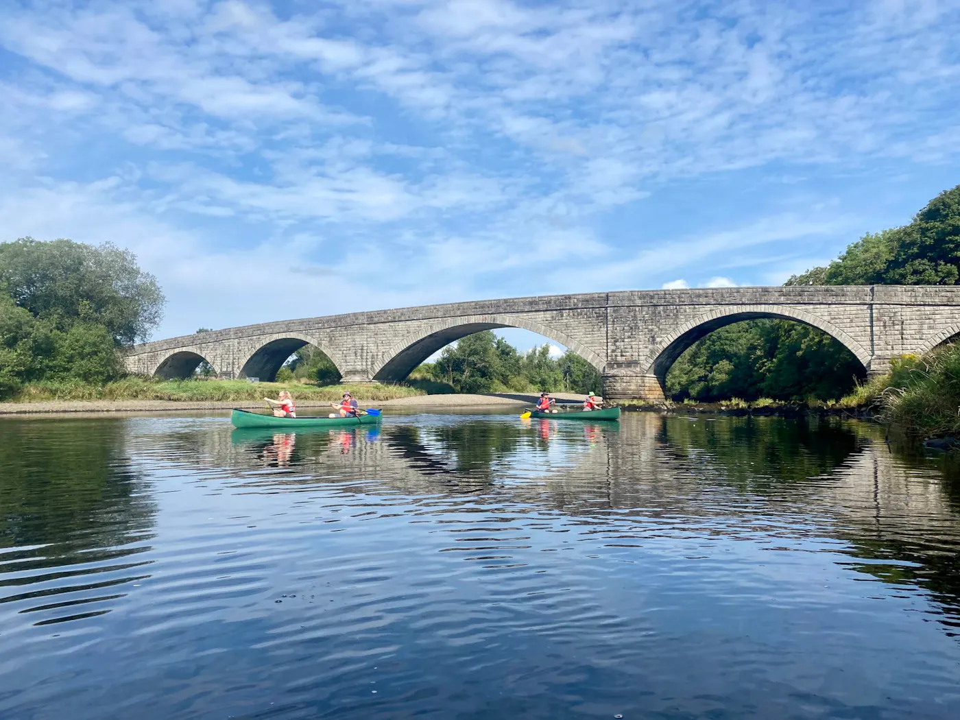 Paddlers in two open canoes on a calm river below an old stone arched bridge