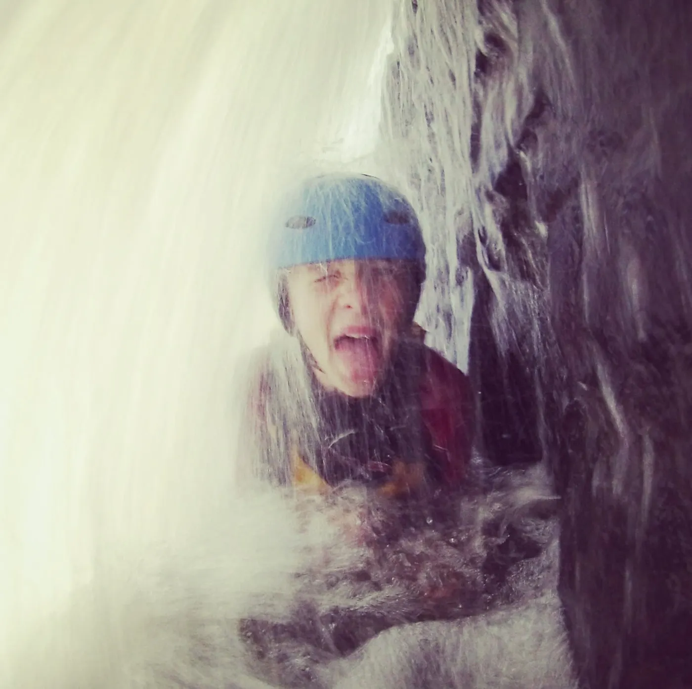 A gorge walker in a blue helmet laughing behind a curtain of falling water