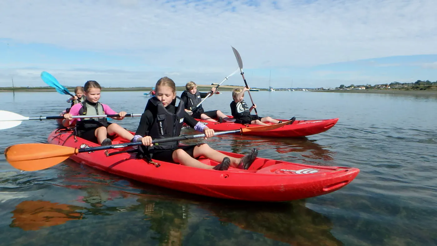 Children paddling red sit-on-top kayaks on calm water at a Mersea Island beach club