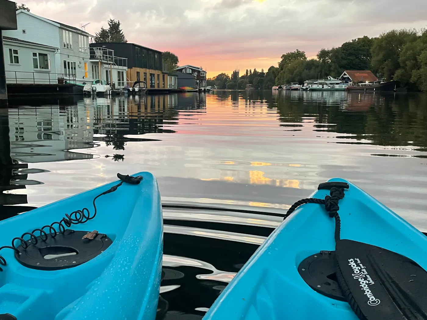 Two blue sit-on-top kayaks on the River Thames at sunset, with houseboats and riverside homes ahead