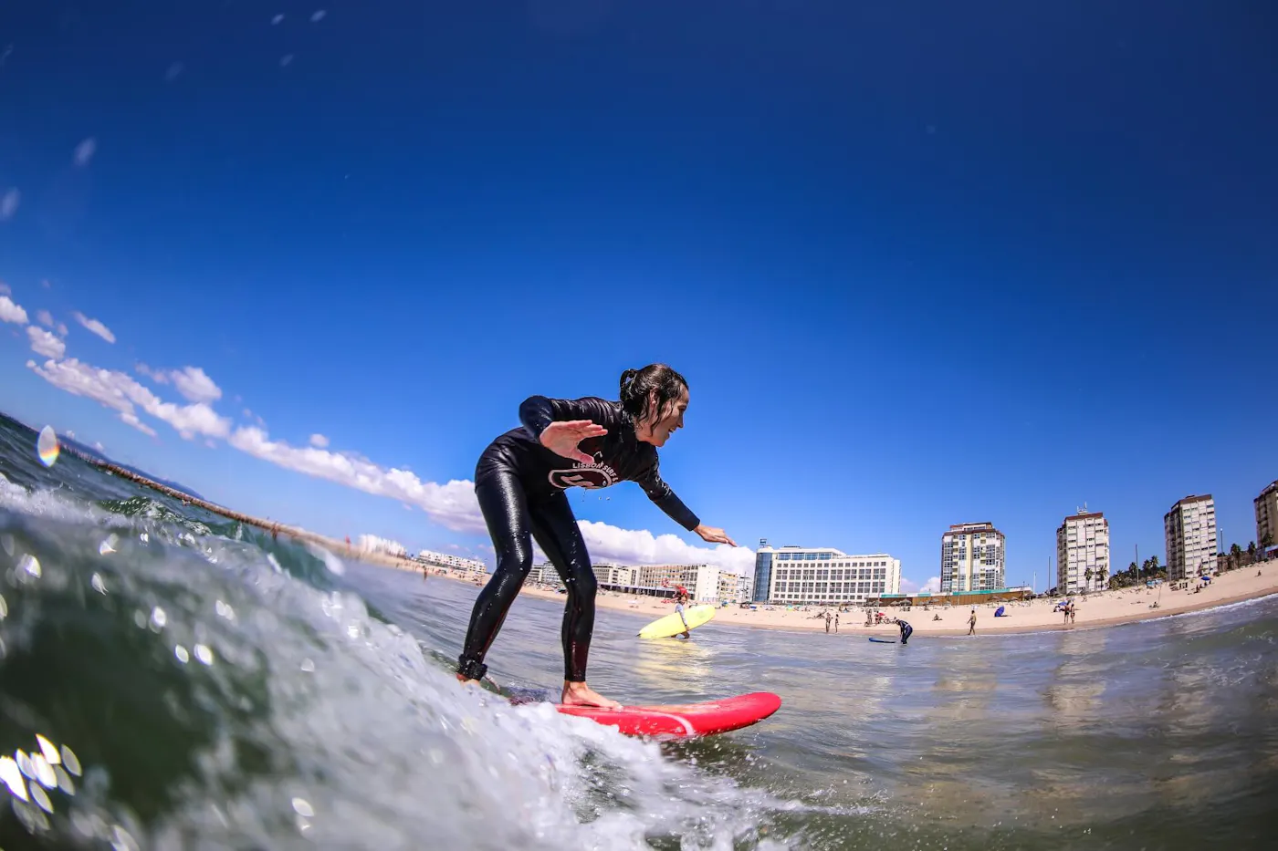A surfer in a wetsuit riding a red board towards the beach and seafront buildings of Costa da Caparica