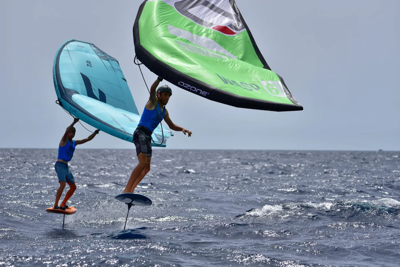Two riders wing foiling above the sea, holding green and blue wings