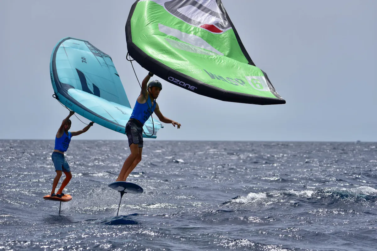 Two riders wing foiling above the sea, holding green and blue wings