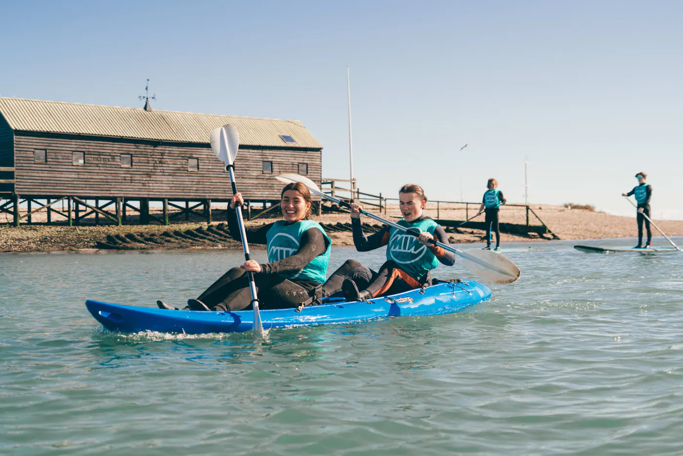 Two young paddlers in a double kayak by the waterfront in West Mersea, with paddleboarders behind