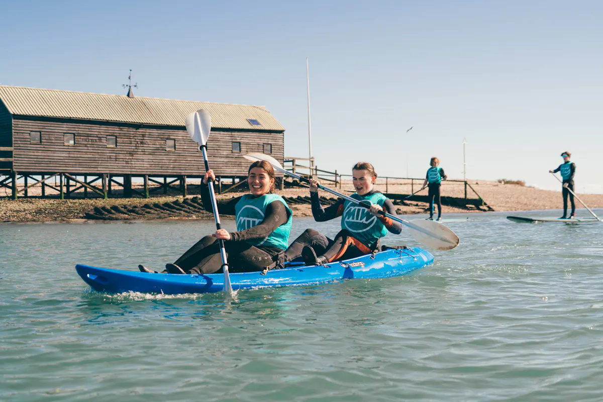 Two young paddlers in a double kayak by the waterfront in West Mersea, with paddleboarders behind