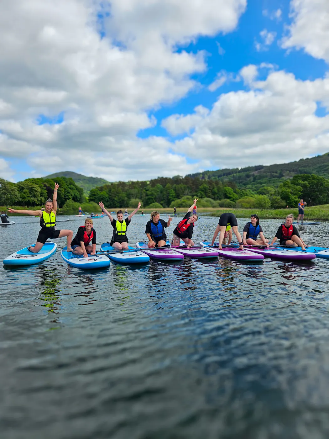 A group of women kneeling and posing on paddleboards in a row on the water, with wooded hills behind