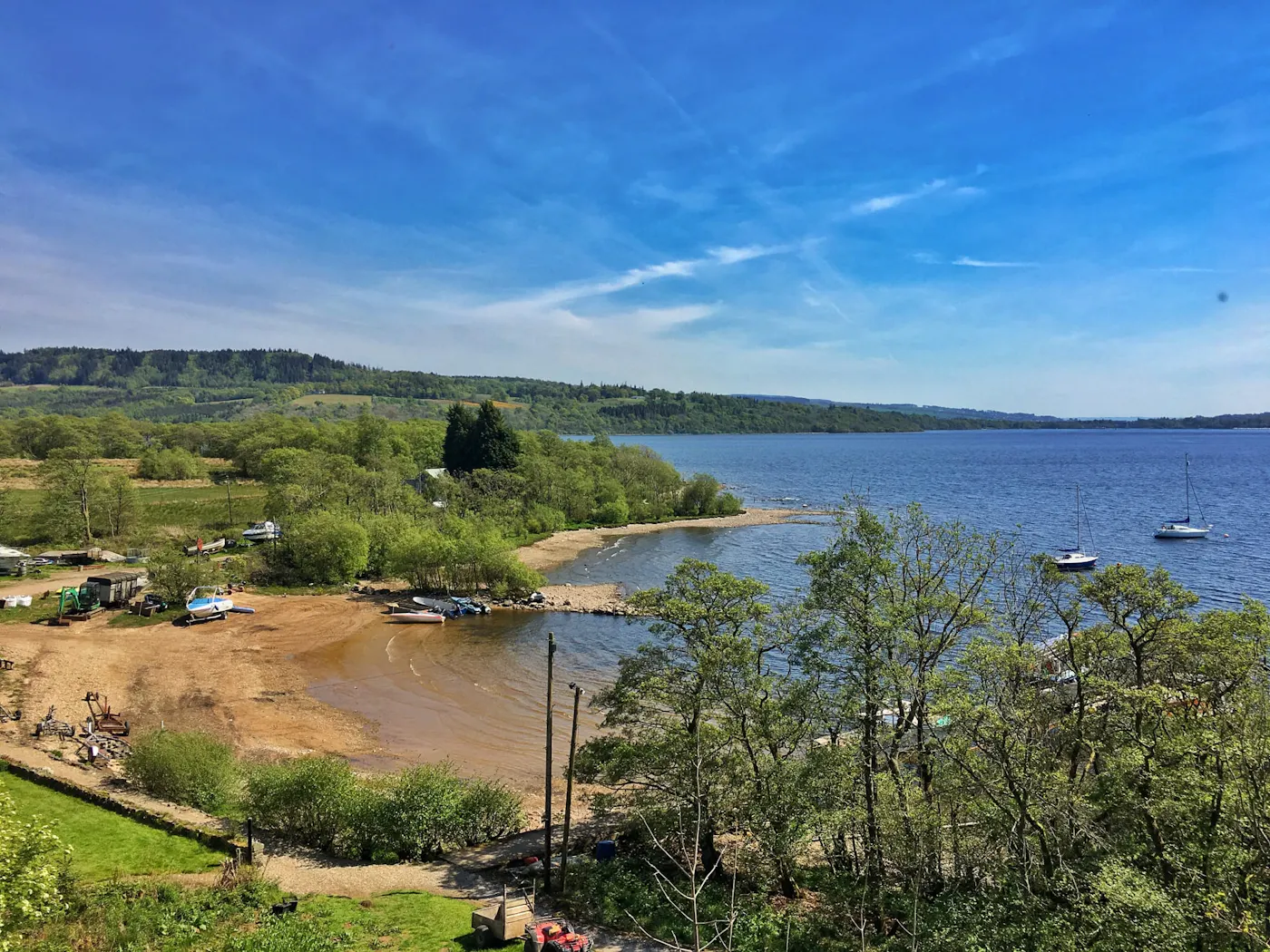 A view over a sandy bay with small boats and moored yachts on Loch Lomond on a sunny day