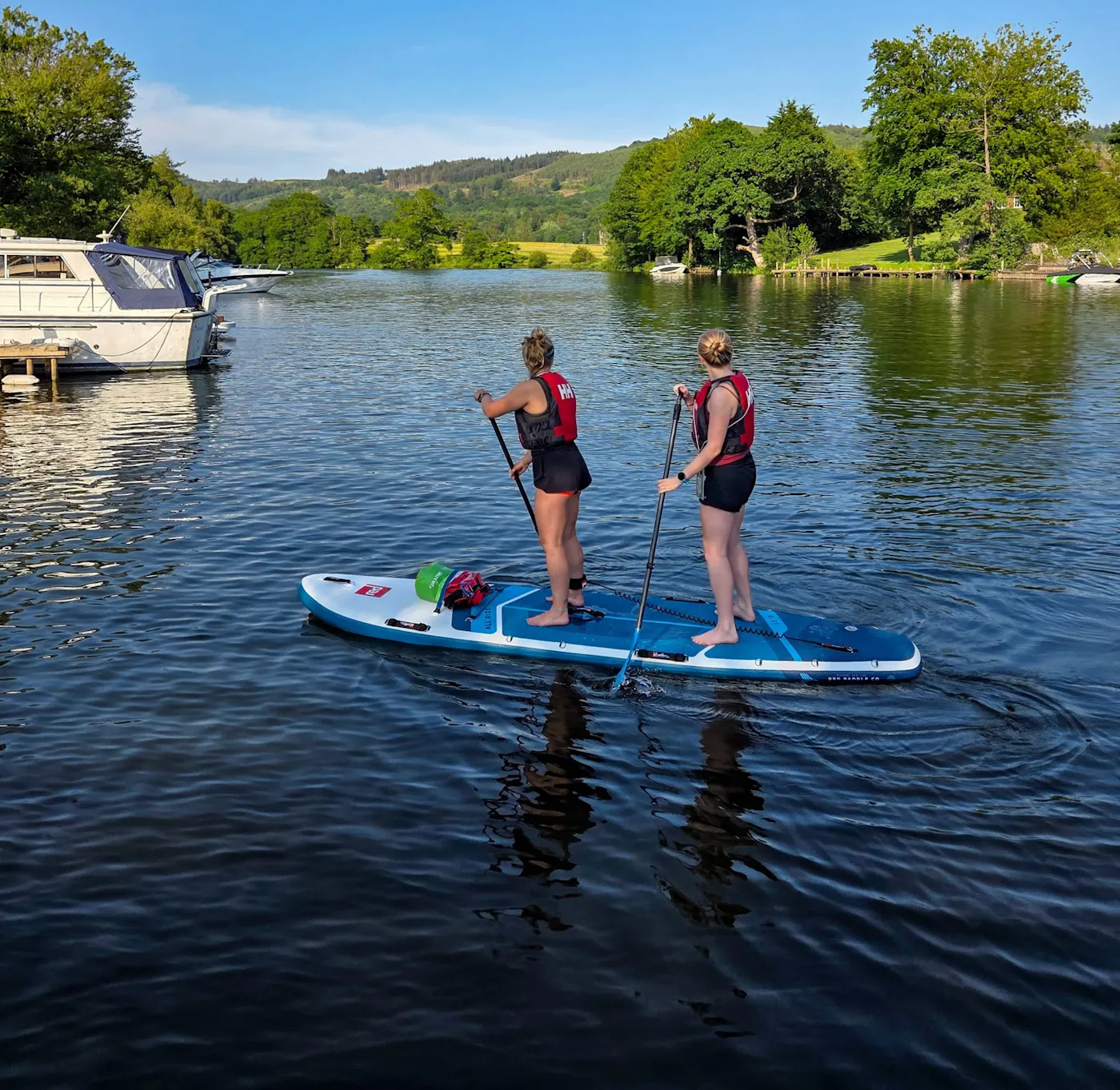 Two people standing together on one large tandem paddleboard, paddling past moored boats