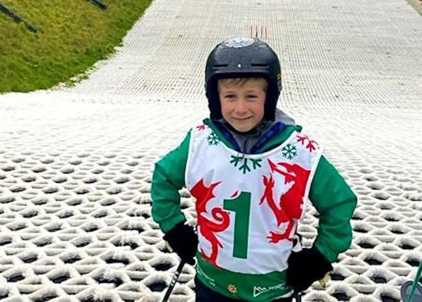 A young skier in a helmet and Welsh race bib standing on a dry ski slope