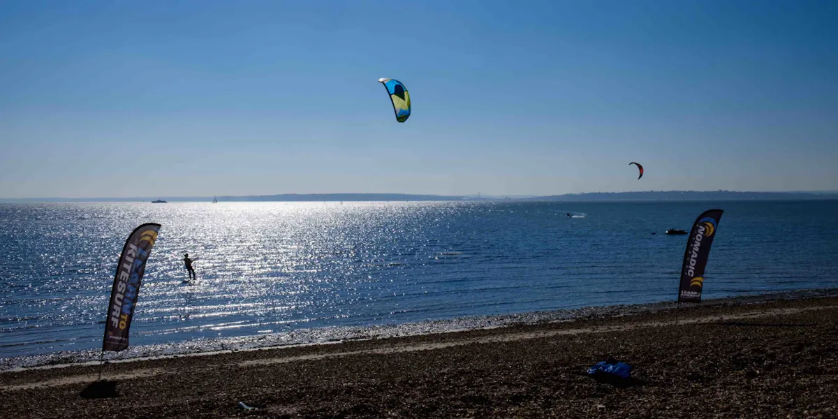 Kitesurfers on the sparkling Solent off Calshot beach, with Nomadic Kitesurf flags on the shingle