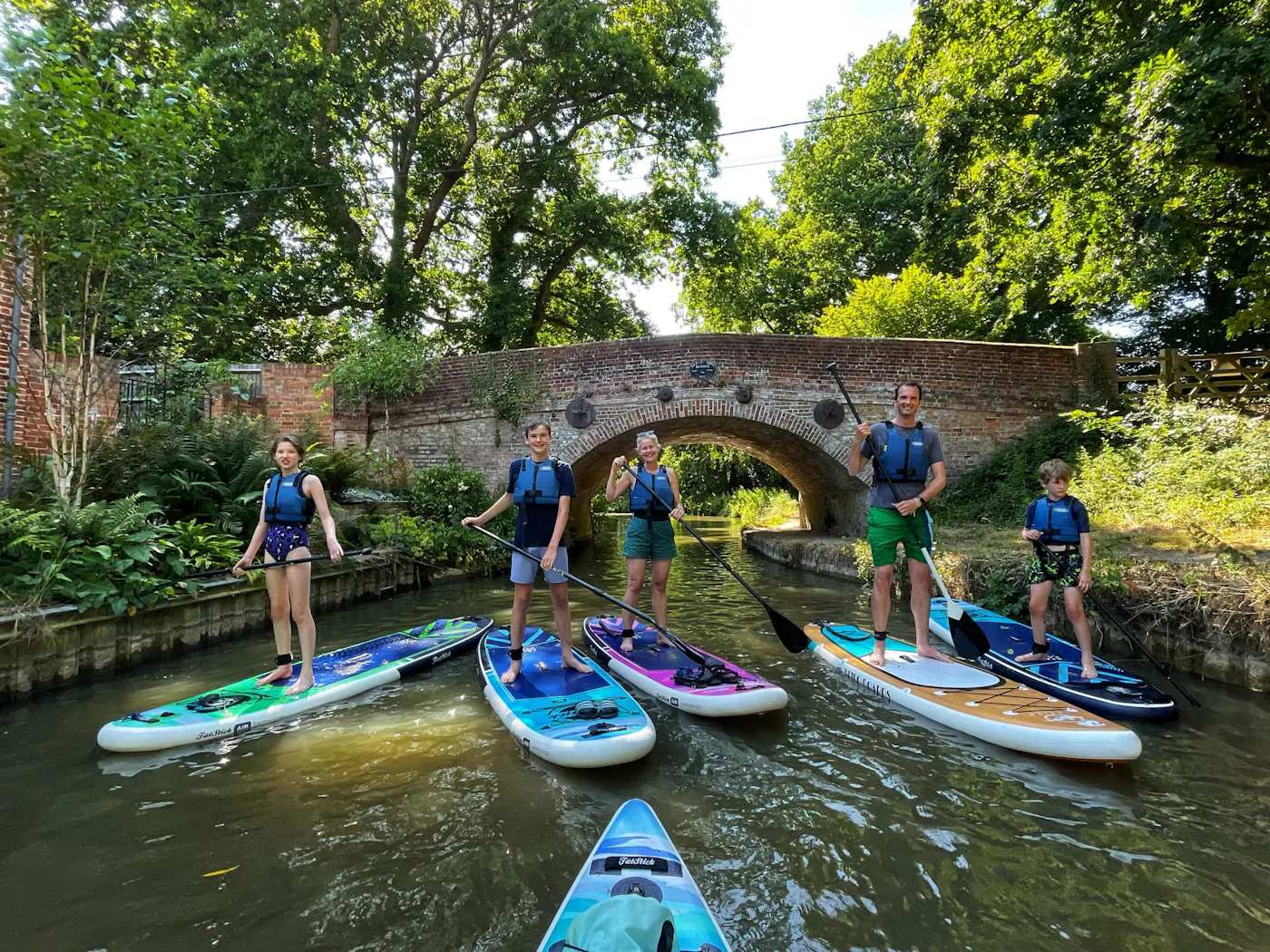A family of paddleboarders standing on their boards beneath a brick bridge on the Basingstoke Canal
