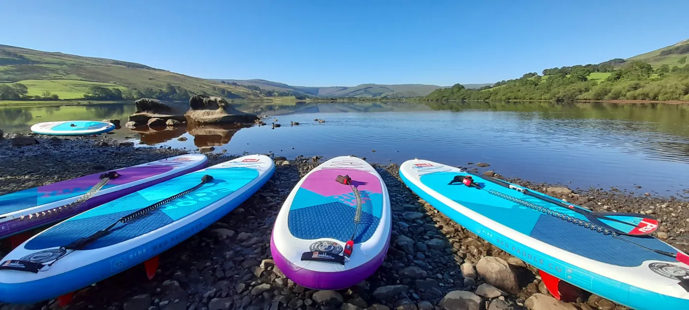 Inflatable paddleboards lined up on a pebbly lake shore beneath green hills on a sunny day