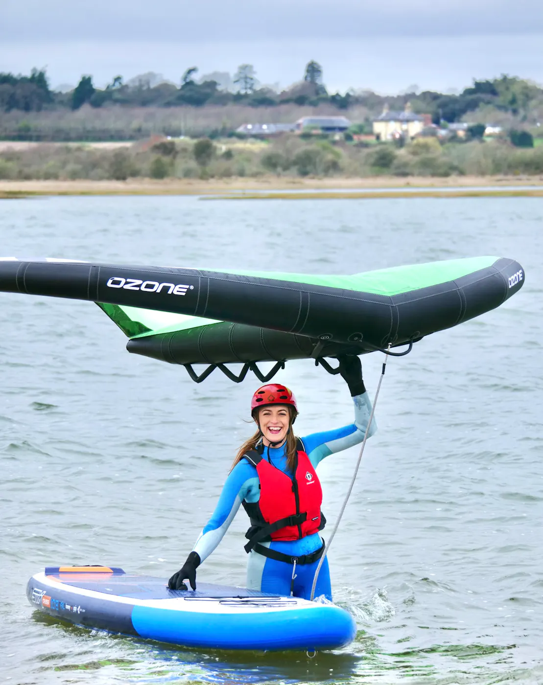 A smiling learner in a helmet and buoyancy aid holding a wing above a paddleboard in a calm lagoon